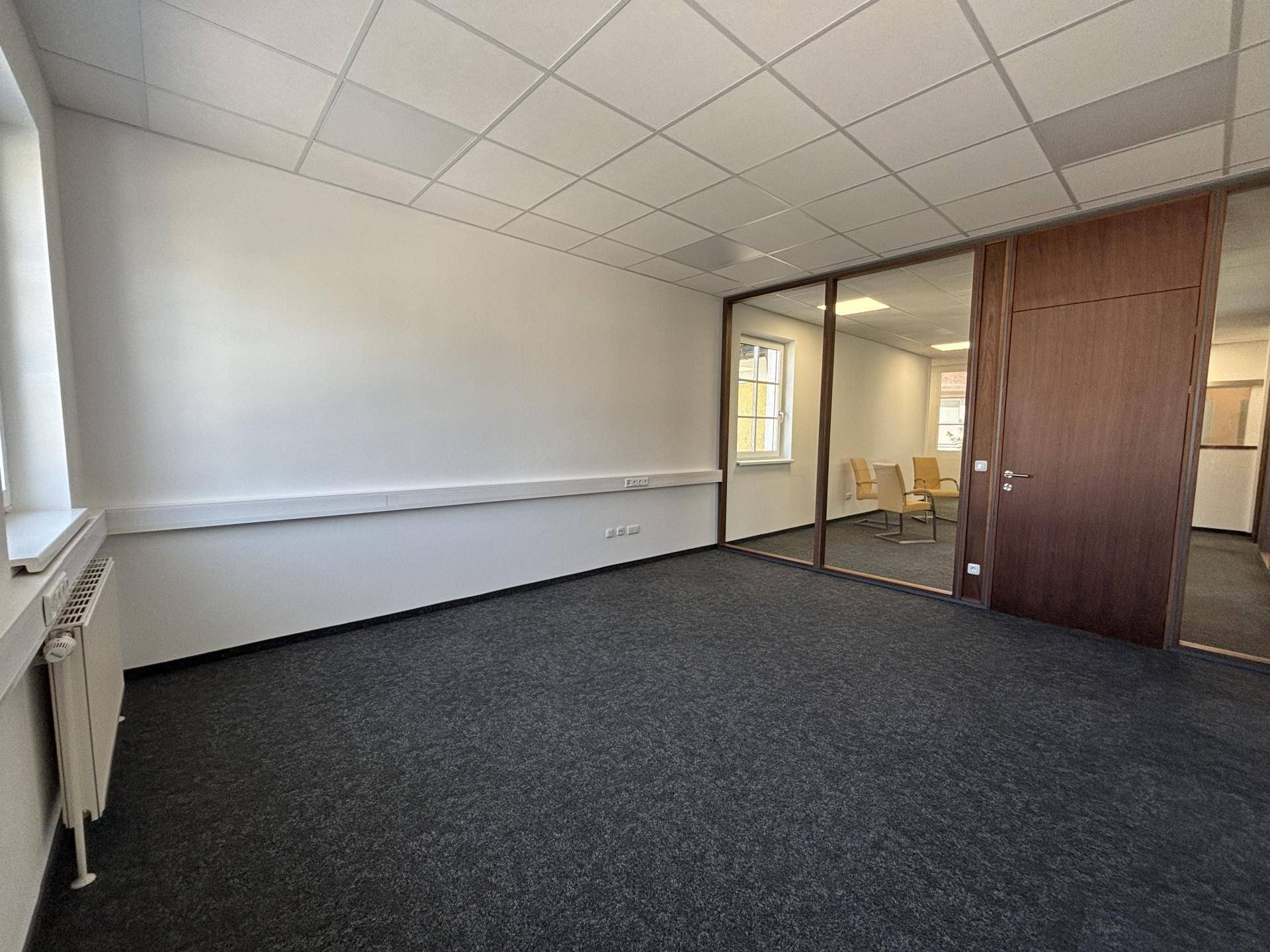 Geräumiges Büro mit Teppichboden und Glaswand zu einem weiteren Arbeitsbereich, gut beleuchtet.