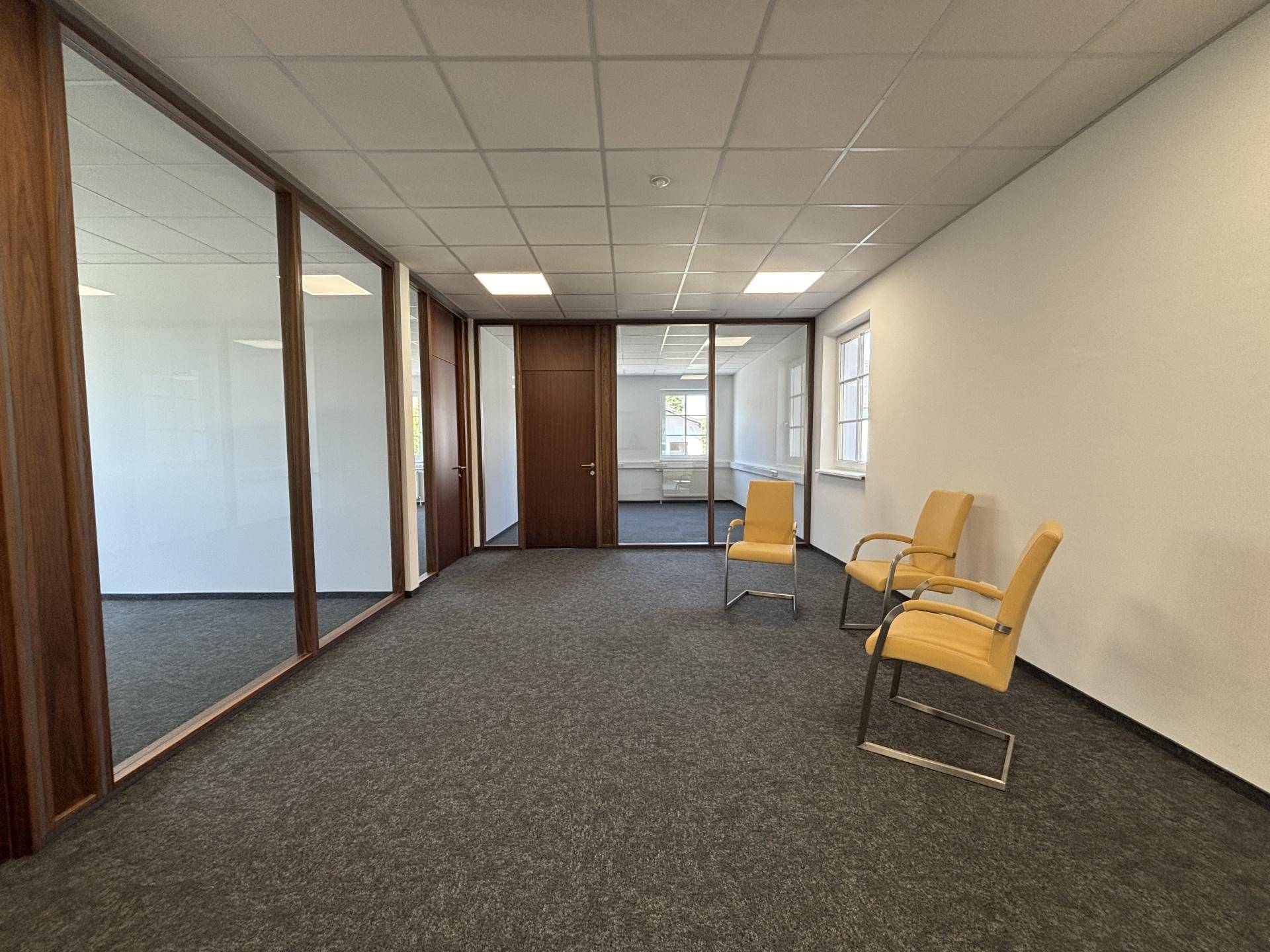 Flur oder Wartebereich in einem Bürogebäude mit Teppichboden, Glaswänden und gelben Stühlen.