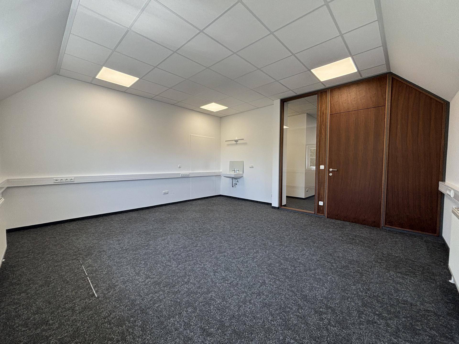 Geräumiger Büroraum mit Teppichboden, abgehängter Decke und integriertem Waschbecken in der Ecke.
