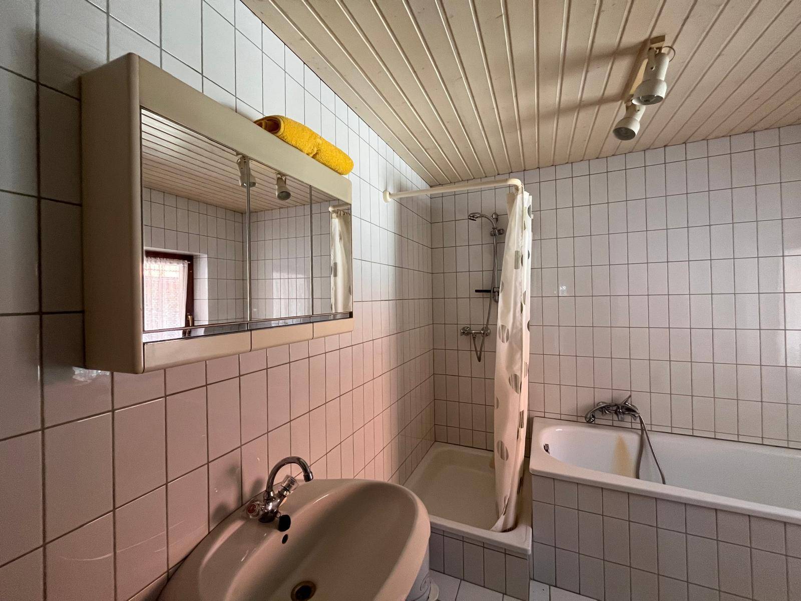 Badezimmer mit weißen Fliesen, Waschbecken, Duschvorhang und einem Spiegelschrank über dem Waschtisch.