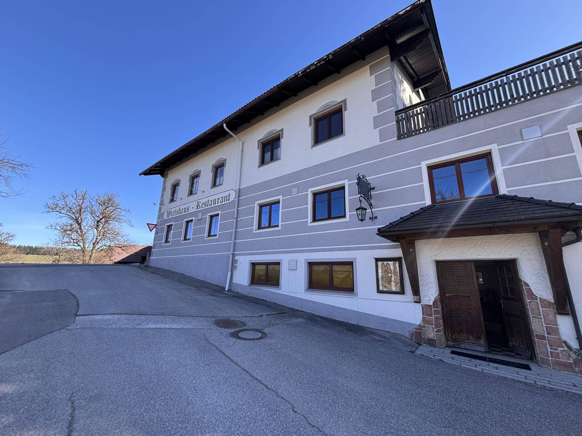 Ansicht des Gebäudes mit Eingangsbereich und Balkon, umgeben von einer gepflasterten Zufahrt.