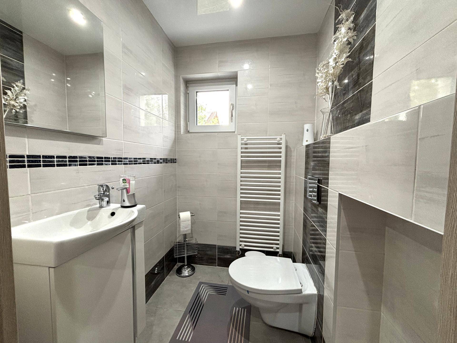 Modernes Gäste-WC mit grauen Fliesen, wandhängendem WC, Waschbecken und Fenster.