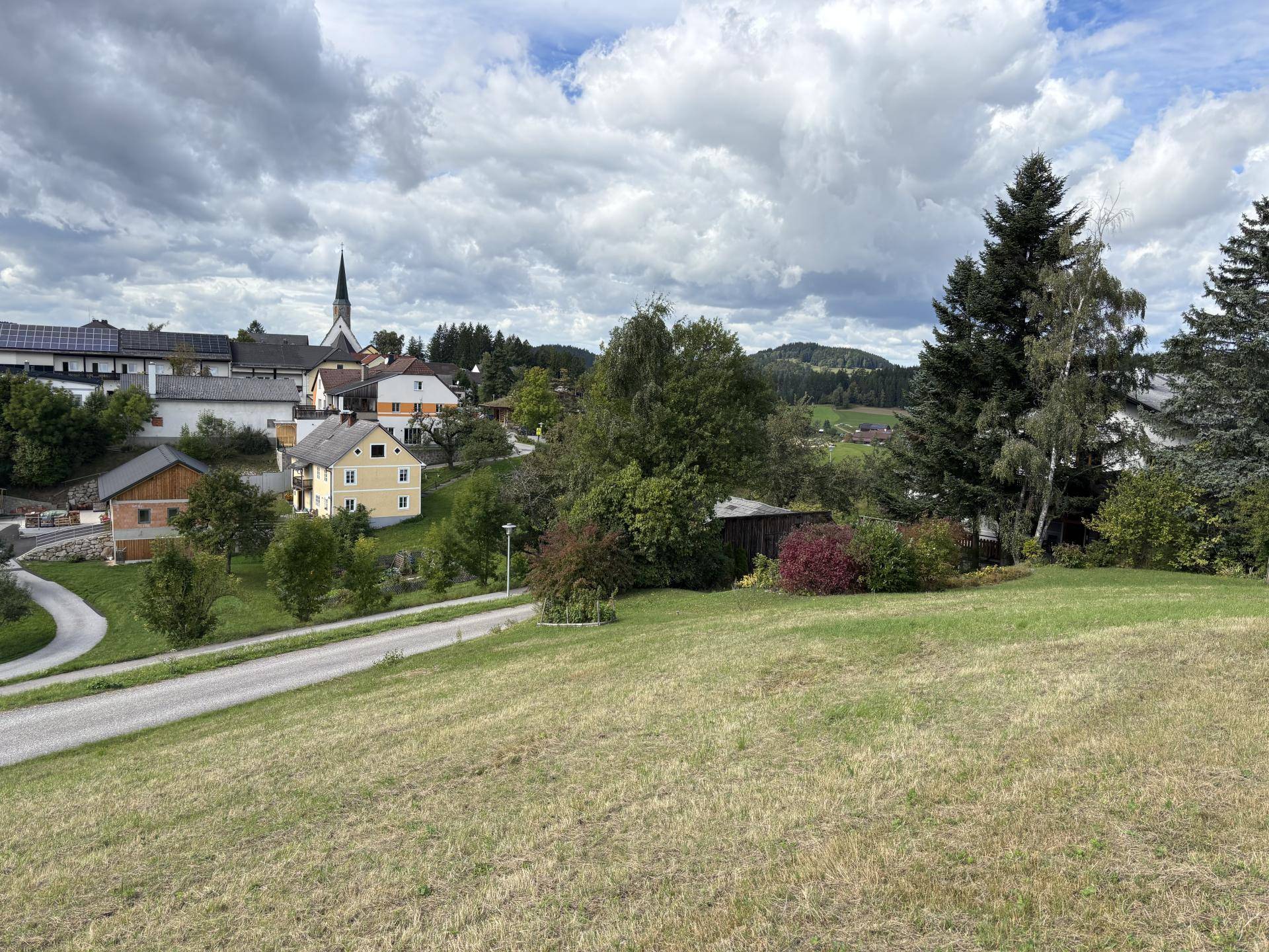 Weitläufiger Blick auf eine ländliche Siedlung mit Kirche und grünen Hügeln unter bewölktem Himmel.