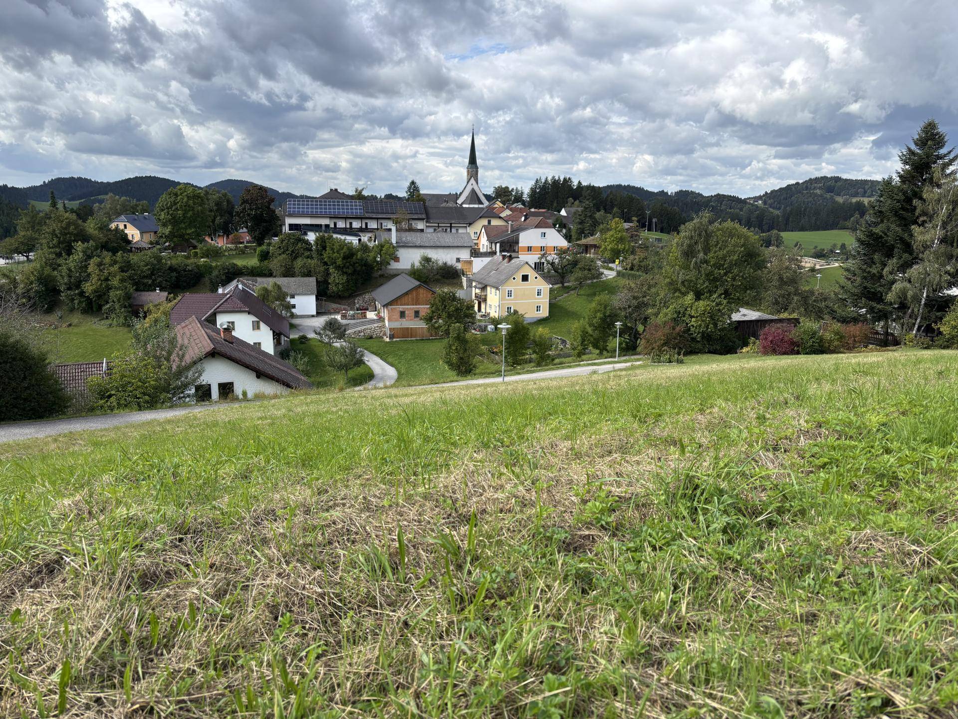 Panoramablick auf ein Dorf mit Kirche und umliegender Naturlandschaft unter einem bewölkten Himmel.
