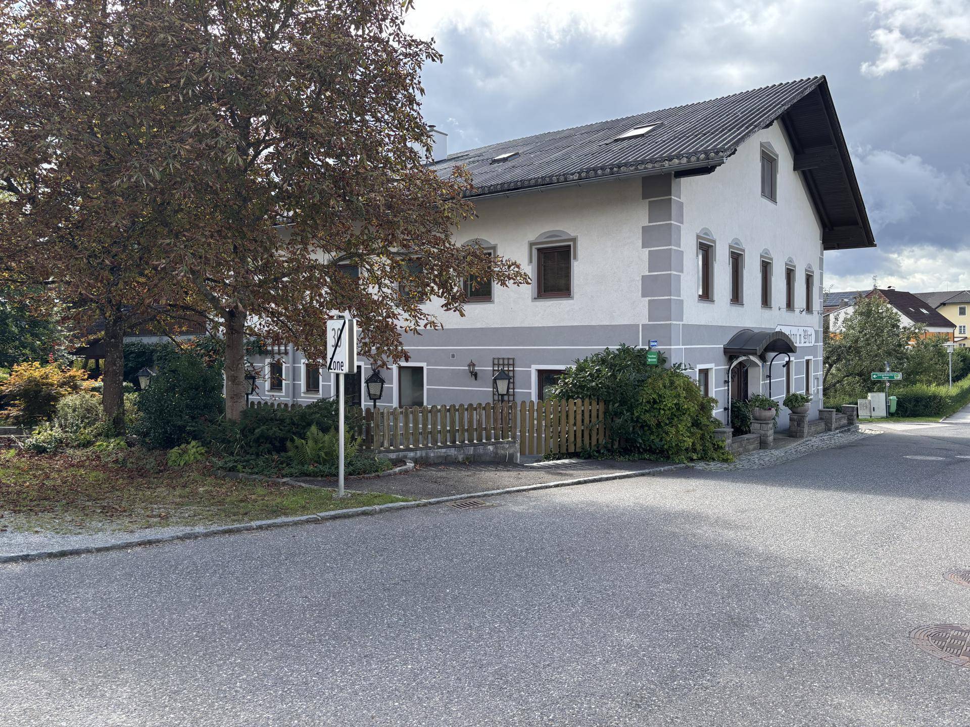 Traditionelles Gebäude mit weiß-grauer Fassade und Satteldach, umgeben von Bäumen und einer Straße.