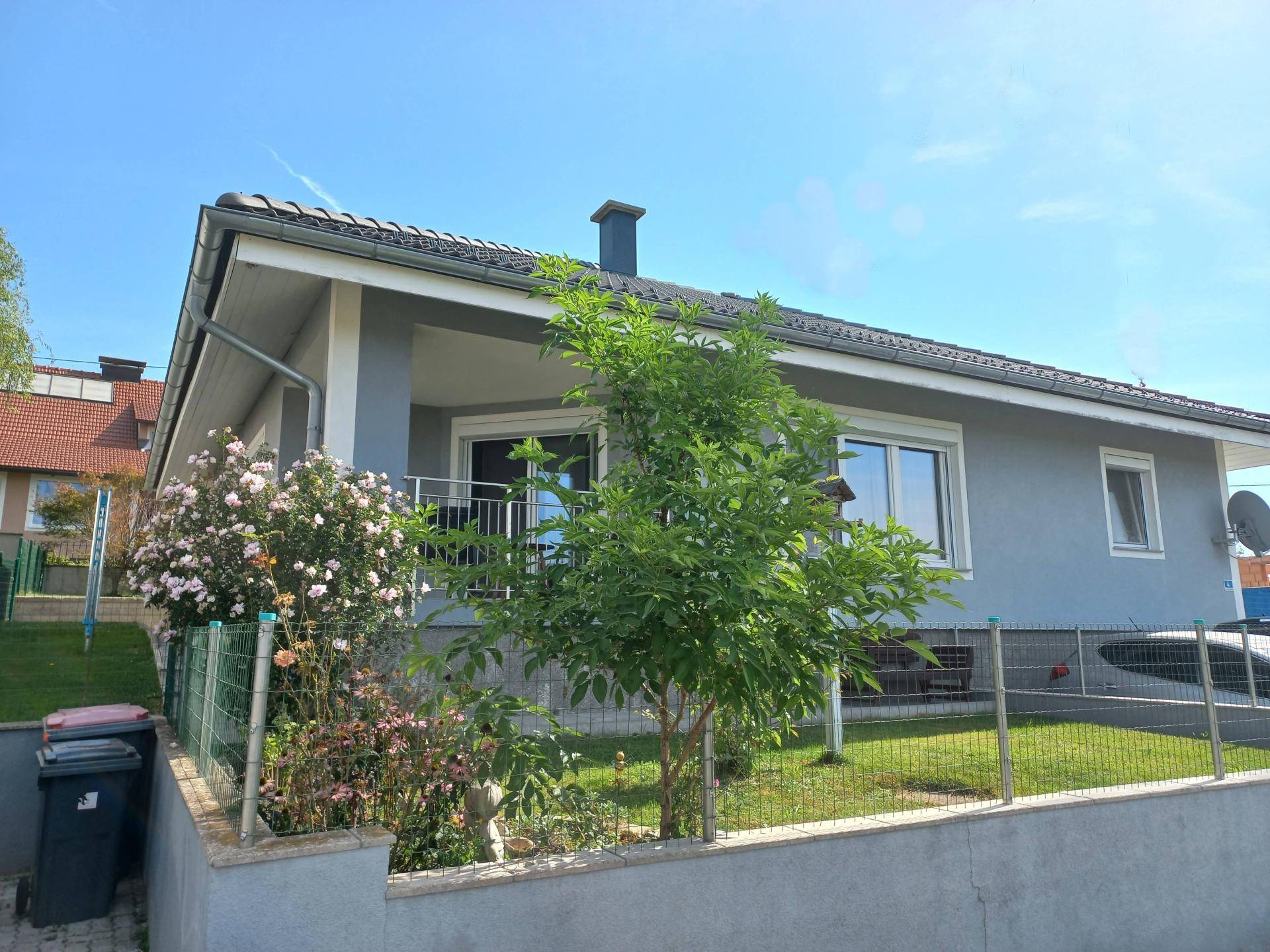 Modernes Einfamilienhaus mit gepflegtem Vorgarten und blühenden Sträuchern unter blauem Himmel.