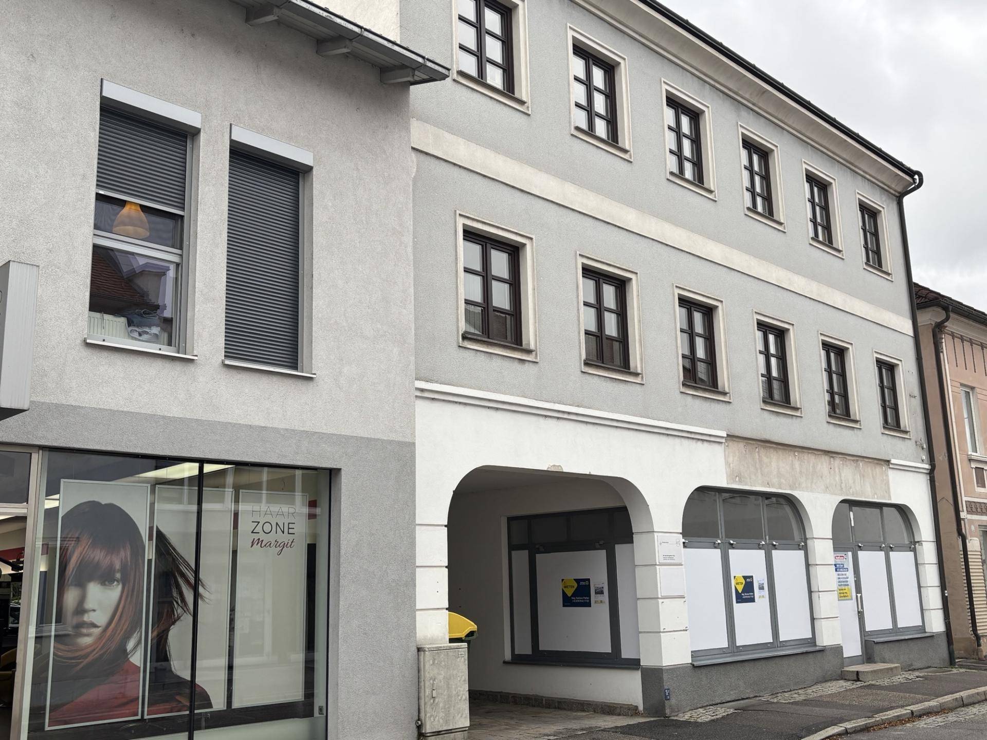Ansicht der Gebäudefassade mit Geschäftsräumen im Erdgeschoss und Fenstern mit Rollläden am angrenzenden Gebäude.