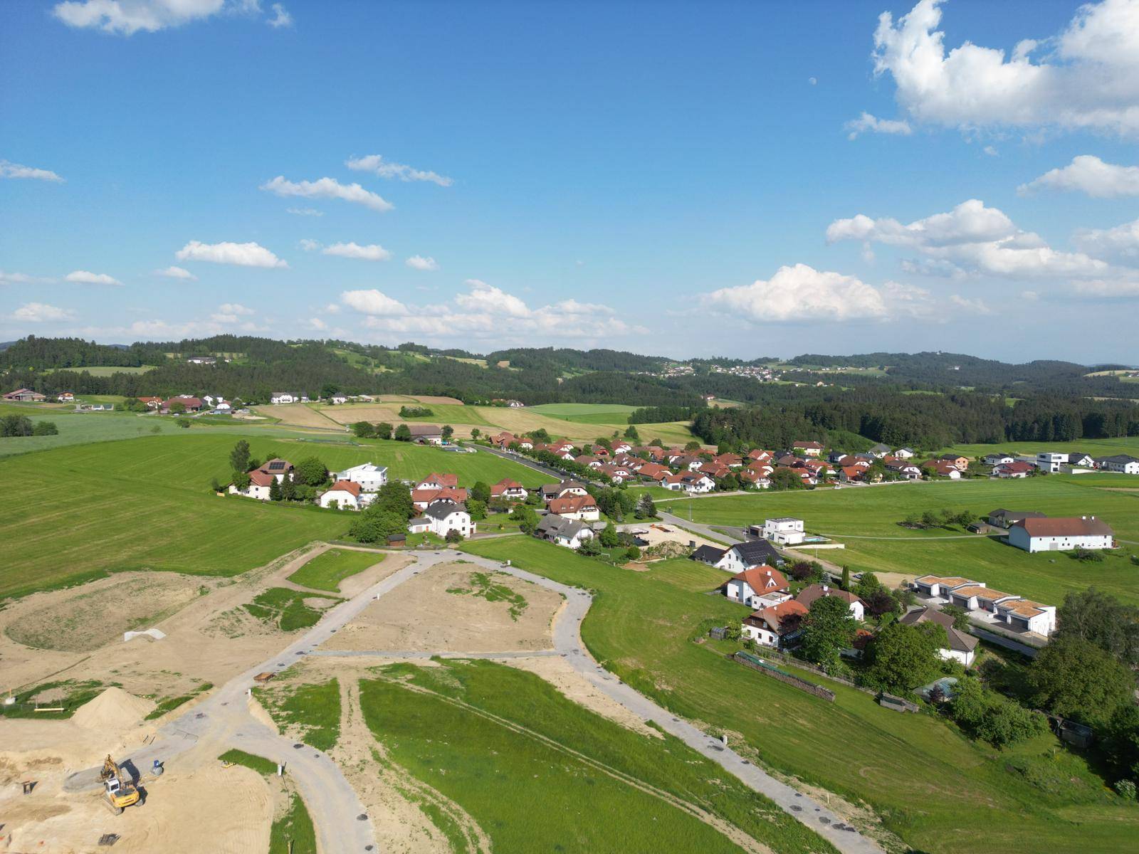 Weitläufige Luftaufnahme einer ländlichen Siedlung eingebettet in grüne Hügel und Felder unter blauem Himmel.