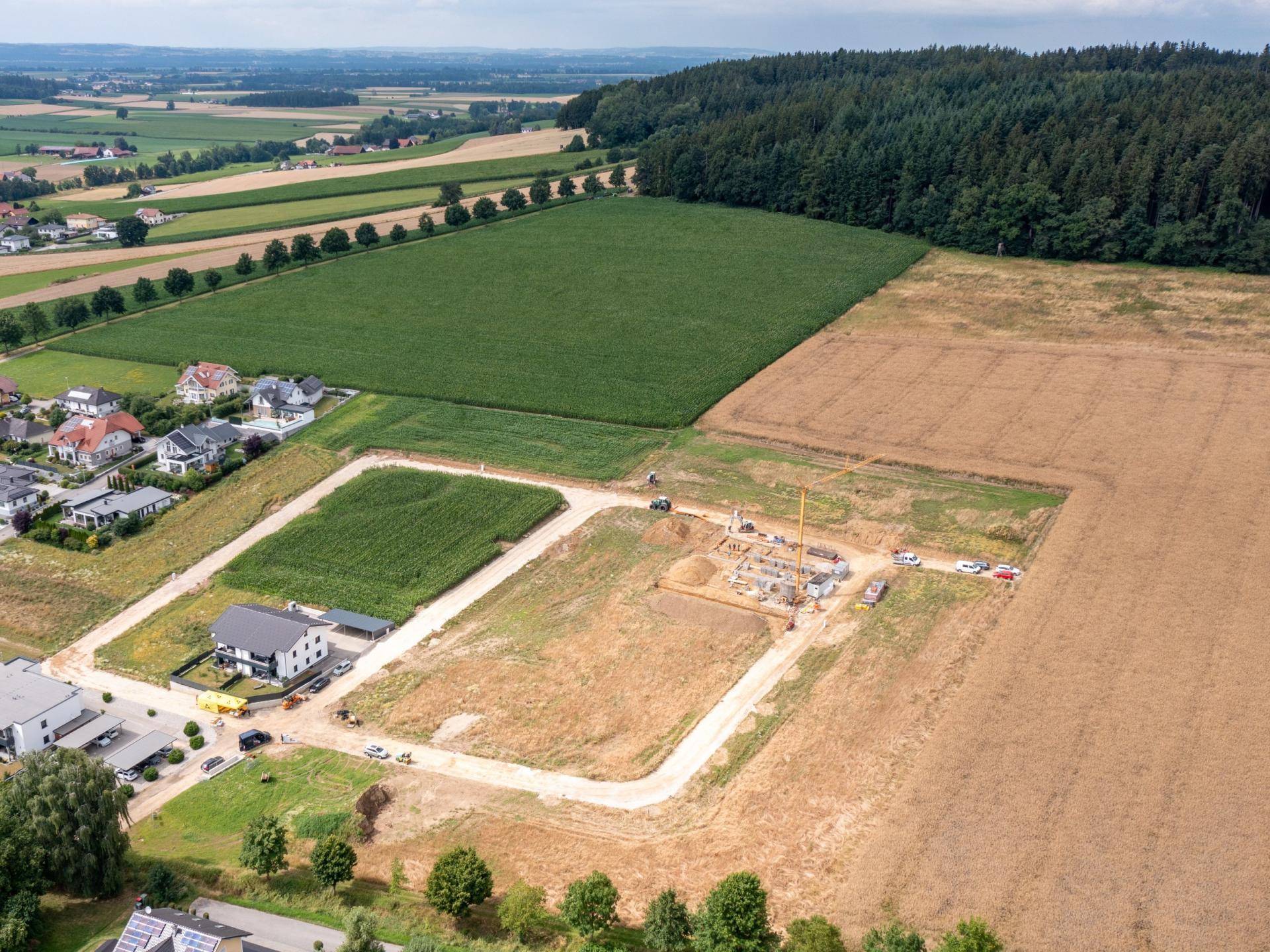 Luftaufnahme eines Neubaugebietes mit aktiver Baustelle, umgeben von Feldern und Wald.