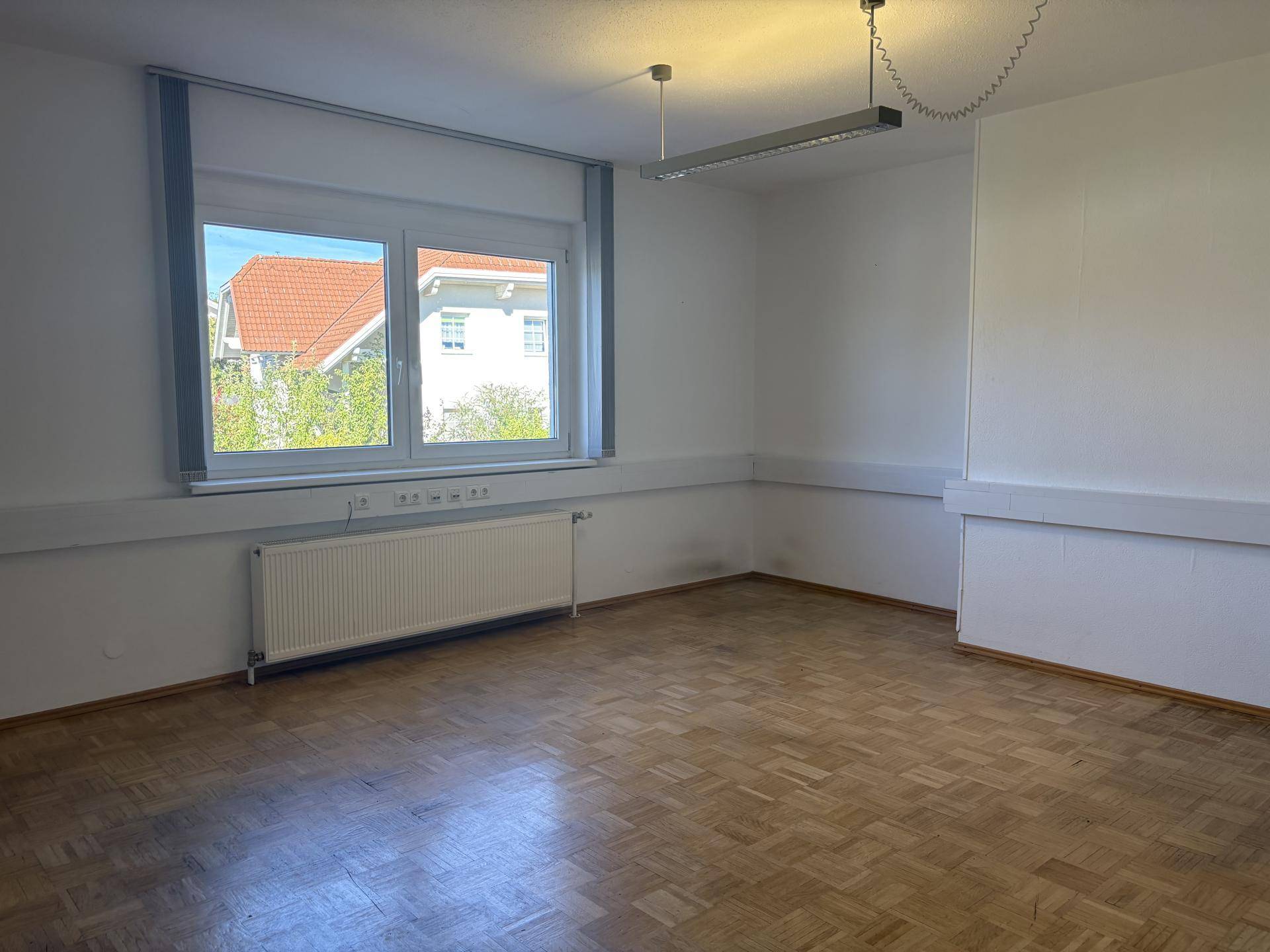 Leerer Büroraum mit Parkettboden und großem Fenster, bietet viel Tageslicht und Ausblick.