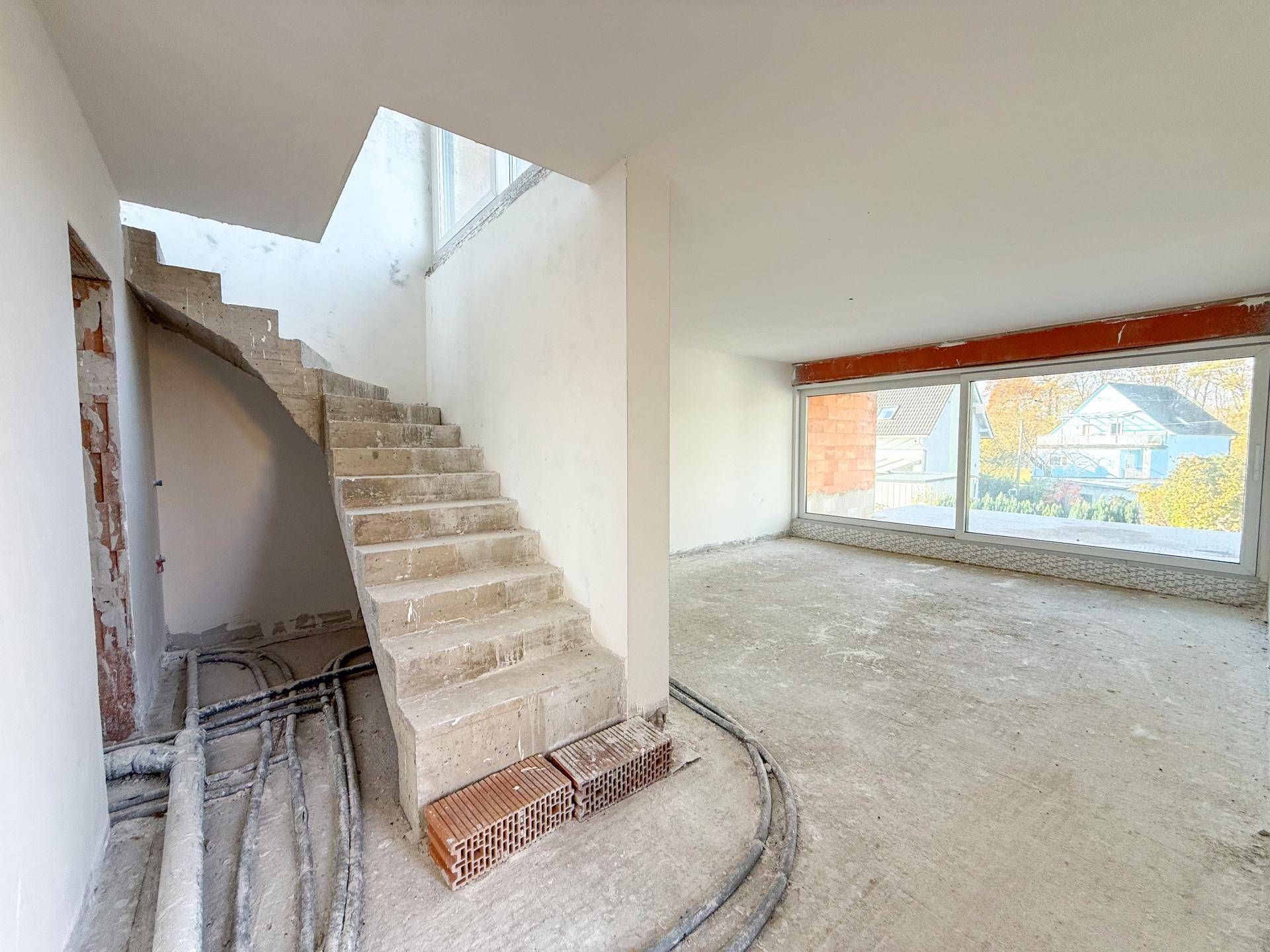 Offener Rohbau-Bereich mit Betontreppe und großen Fenstern, die viel Licht hereinlassen.