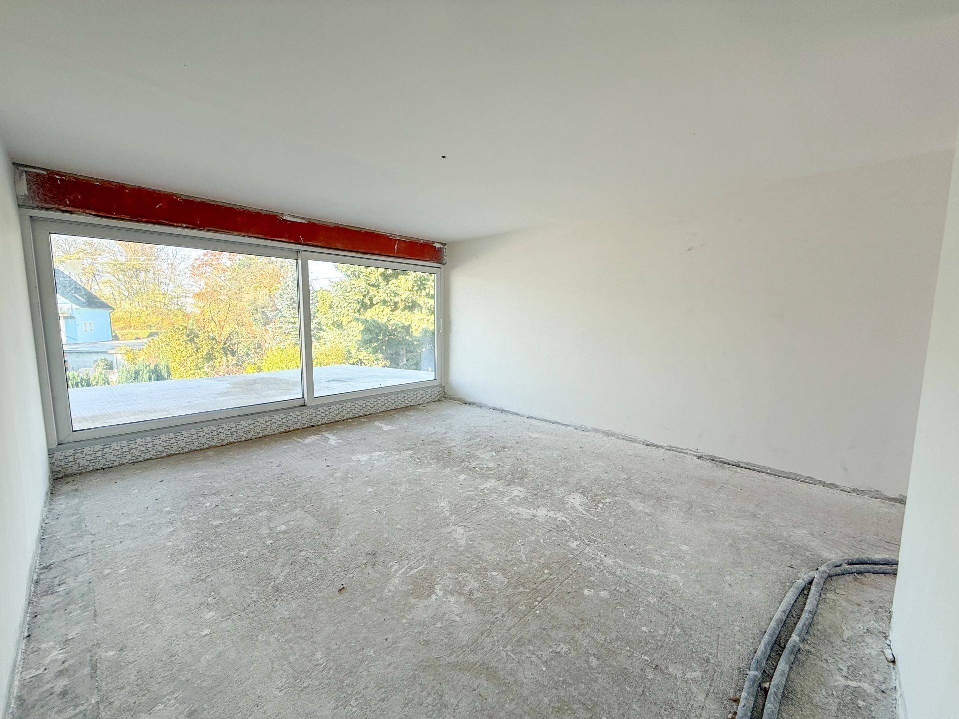 Heller Rohbau-Raum mit großer Fensterfront und Blick auf die umliegende Natur.