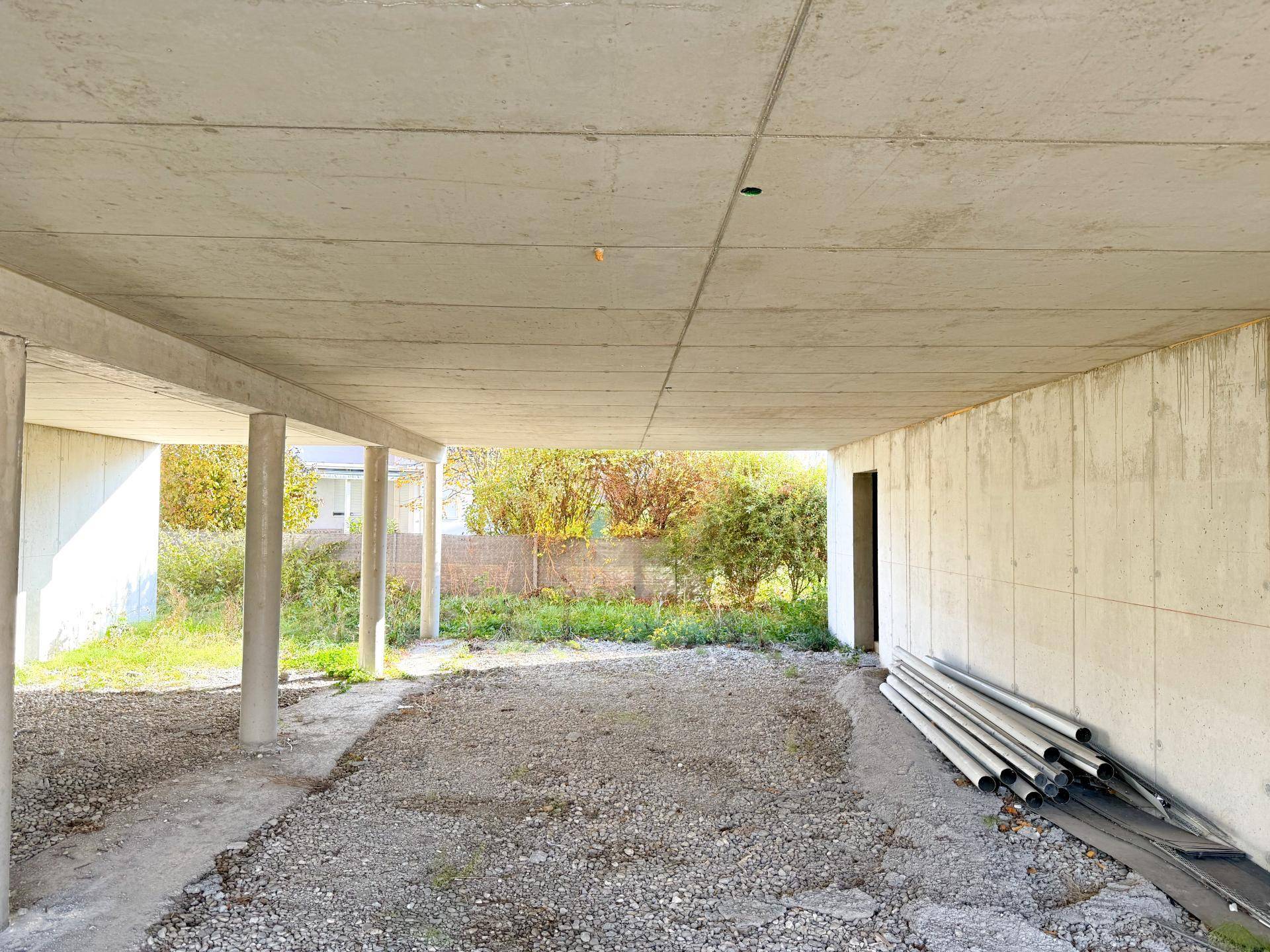 Offener Bereich unter dem Gebäude mit Betonpfeilern und Blick ins Grüne.
