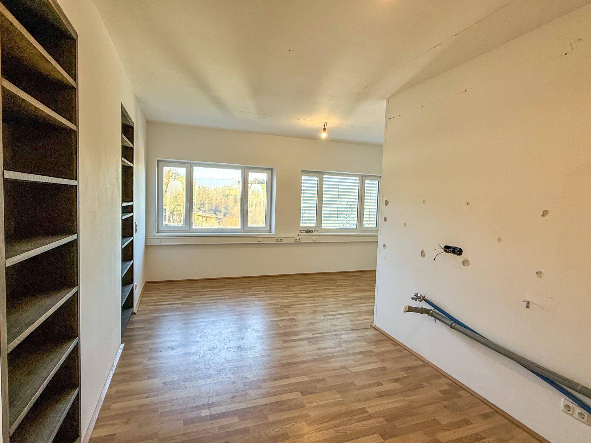 Leerer Raum mit eingebauten Regalen, Fenstern und sichtbaren Installationen, die auf Renovierungsbedarf hinweisen.