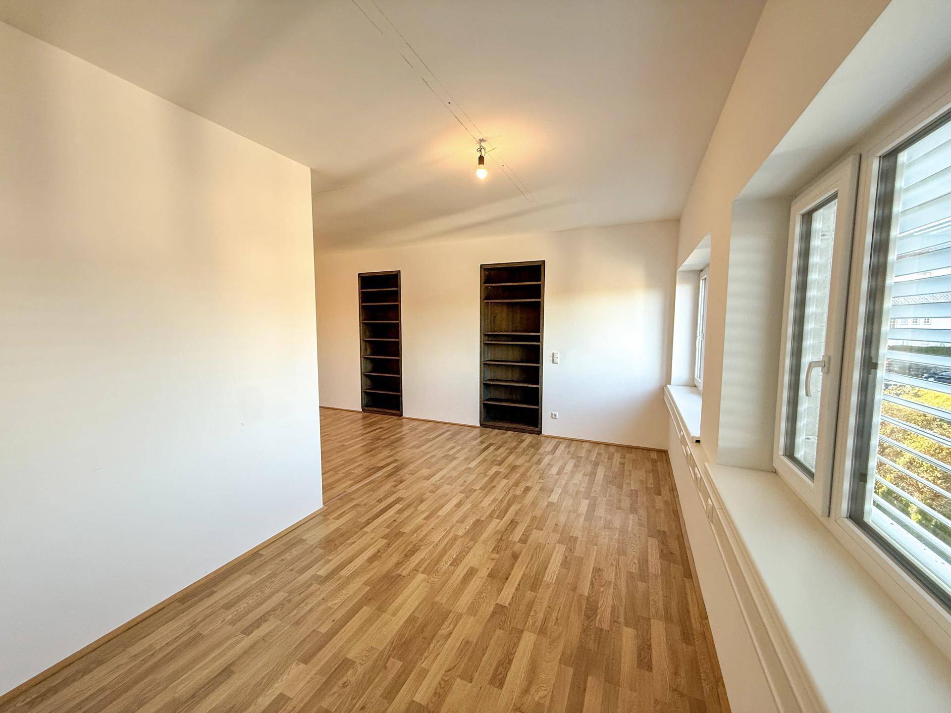Heller Raum mit Holzboden, eingebauten Regalen und Fenstern, die Tageslicht hereinlassen.