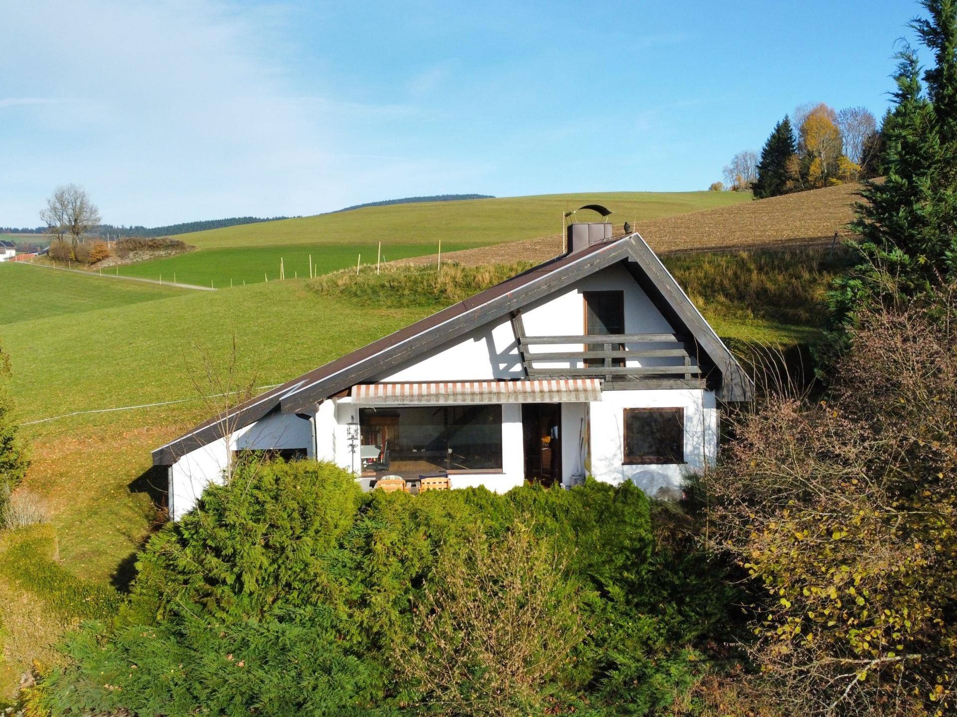 Haus in ländlicher Umgebung mit grünen Hügeln und Bäumen unter blauem Himmel.