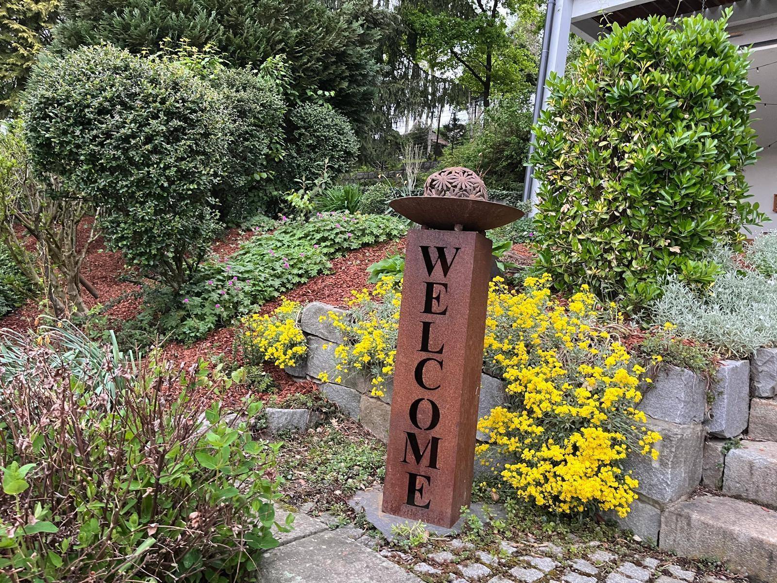 Rostfarbene "WELCOME"-Säule als Gartendekoration, umgeben von gelben Blumen und Steinmauern.