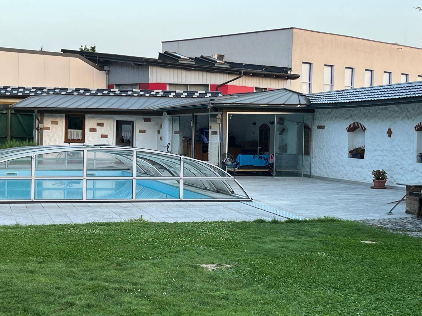 Überdachter Poolbereich mit angrenzender Terrasse und Gebäuden im Hintergrund unter hellem Himmel.