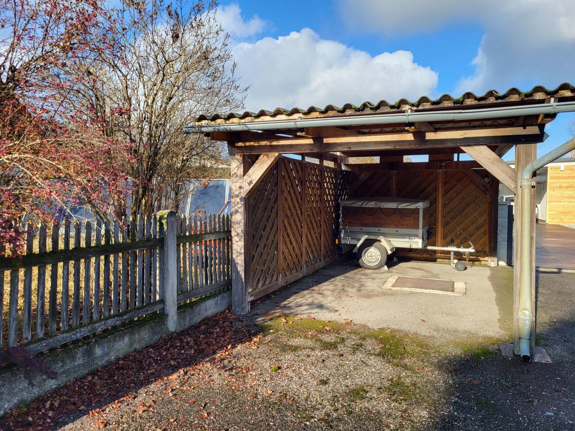 Überdachter Holzcarport mit Zaun und Abstellmöglichkeit für einen Anhänger.