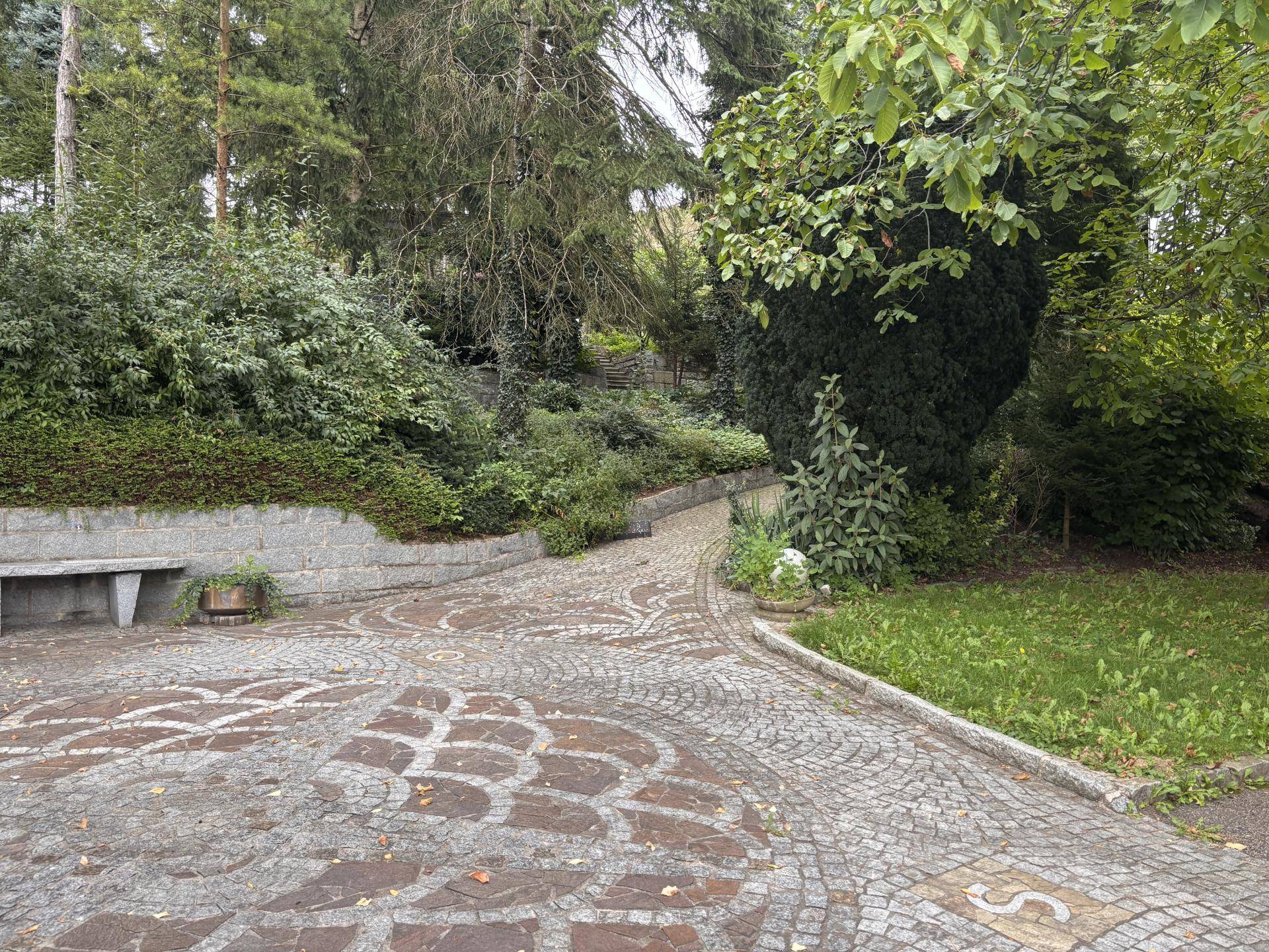 Geschwungener Kopfsteinpflasterweg durch einen grünen Garten mit Bäumen und Sträuchern.