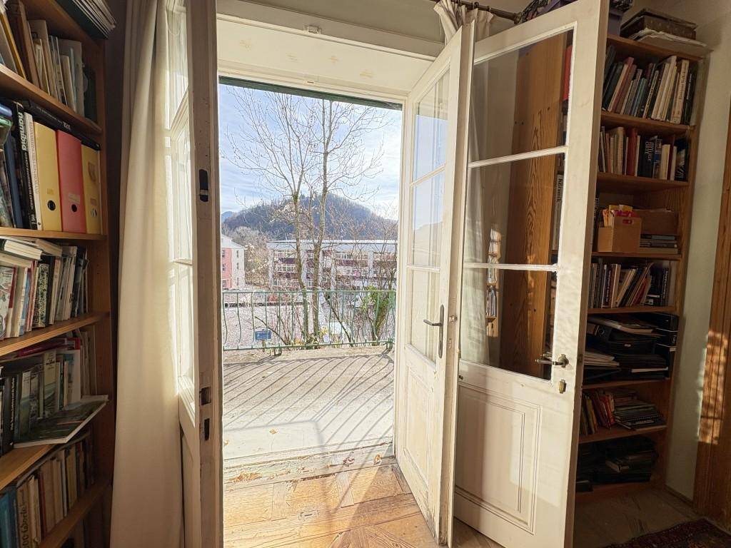 Heller Raum mit Bücherregalen, der sich durch Flügeltüren zu einem Balkon mit Bergblick öffnet.