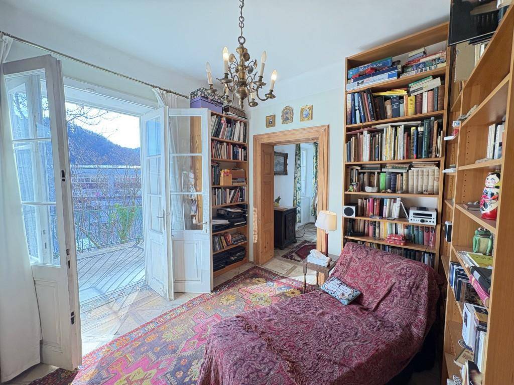Helles Wohnzimmer mit Bücherregalen, Chaiselongue und Zugang zu einem Balkon mit See- und Bergblick.