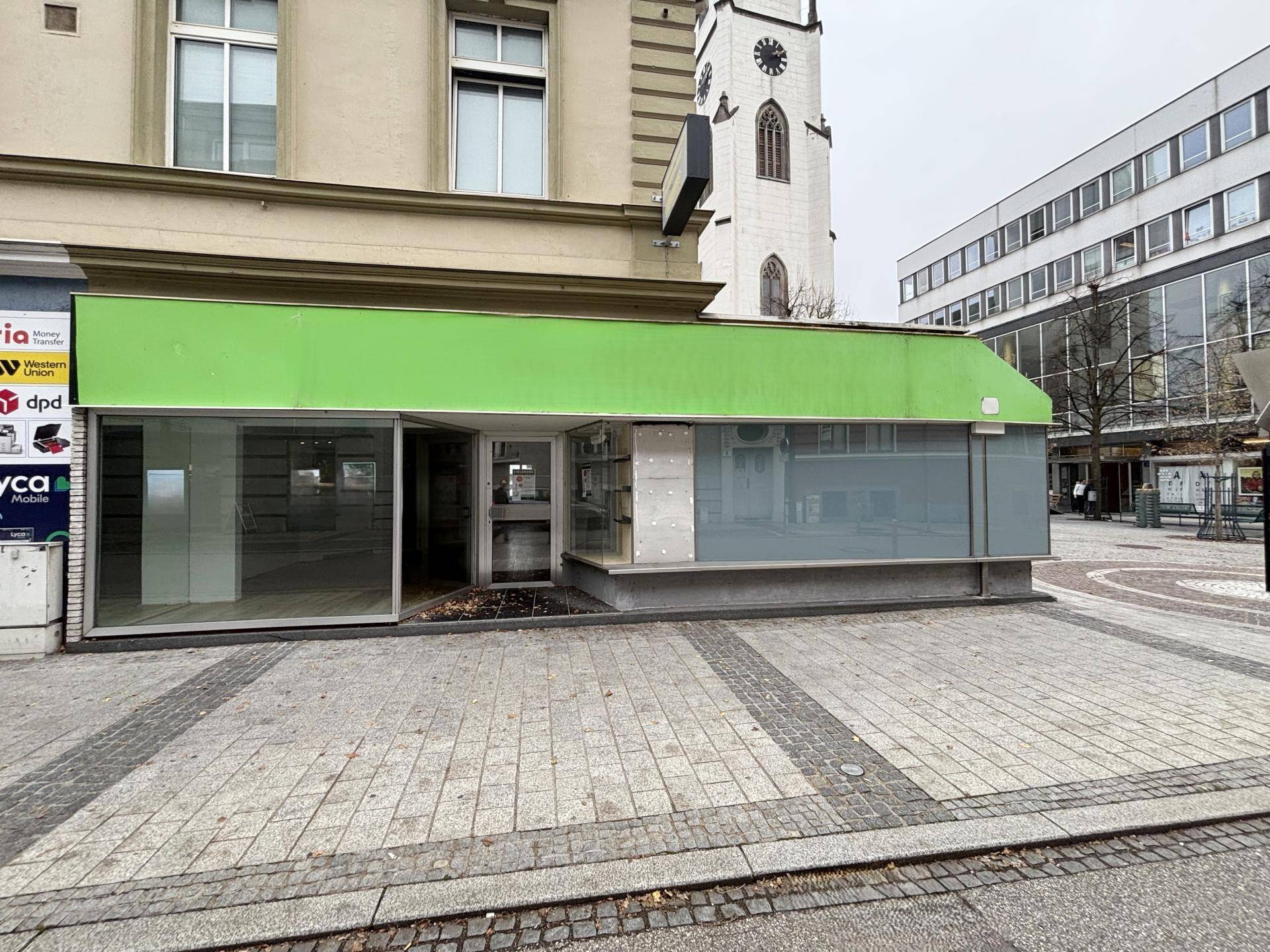 Außenansicht eines Geschäftsgebäudes mit großer Schaufensterfront und grüner Markise an einem Stadtplatz.