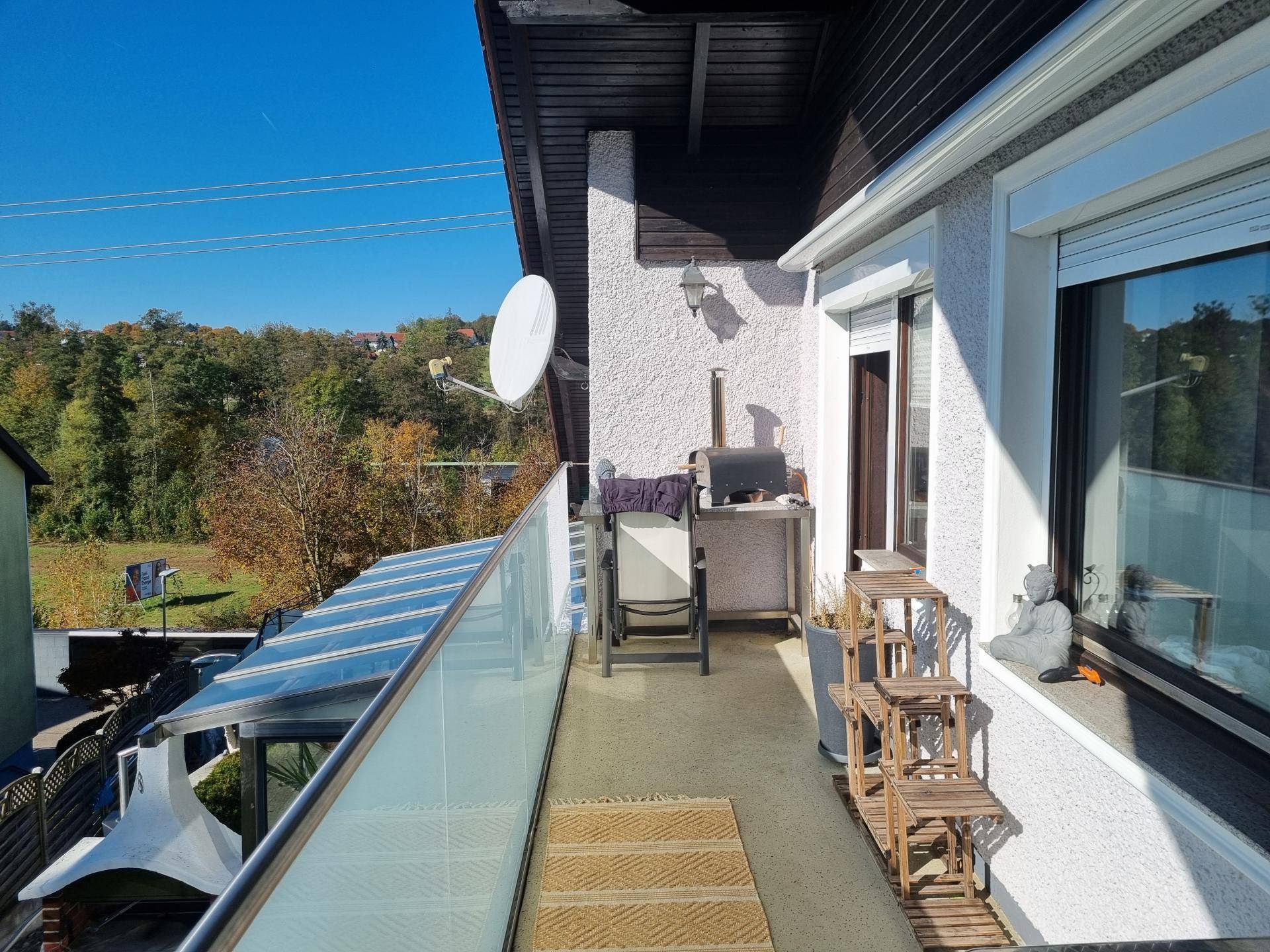 Geräumiger Balkon mit Glasbrüstung, Sitzgelegenheiten und weitem Blick ins Grüne unter blauem Himmel.