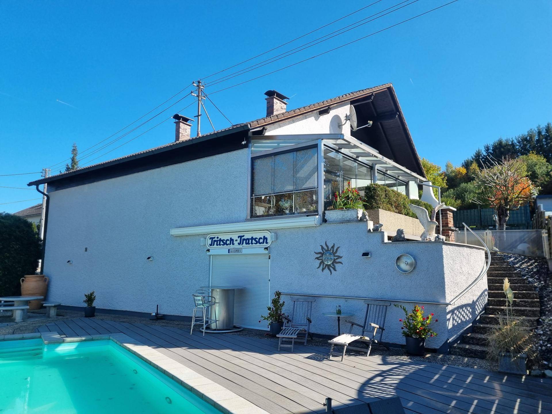 Rückansicht des Hauses mit Pool, Sonnenterrasse und Wintergarten unter blauem Himmel.