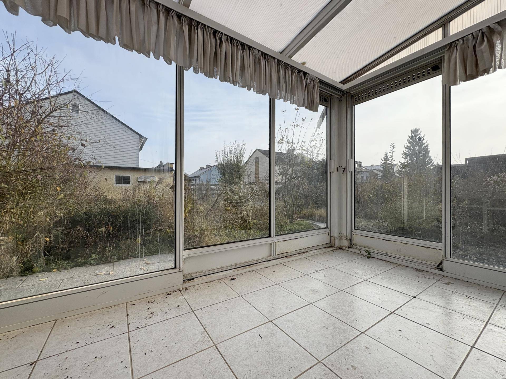 Wintergarten mit Fliesenboden und großen Fenstern, die einen Blick in den Garten bieten.