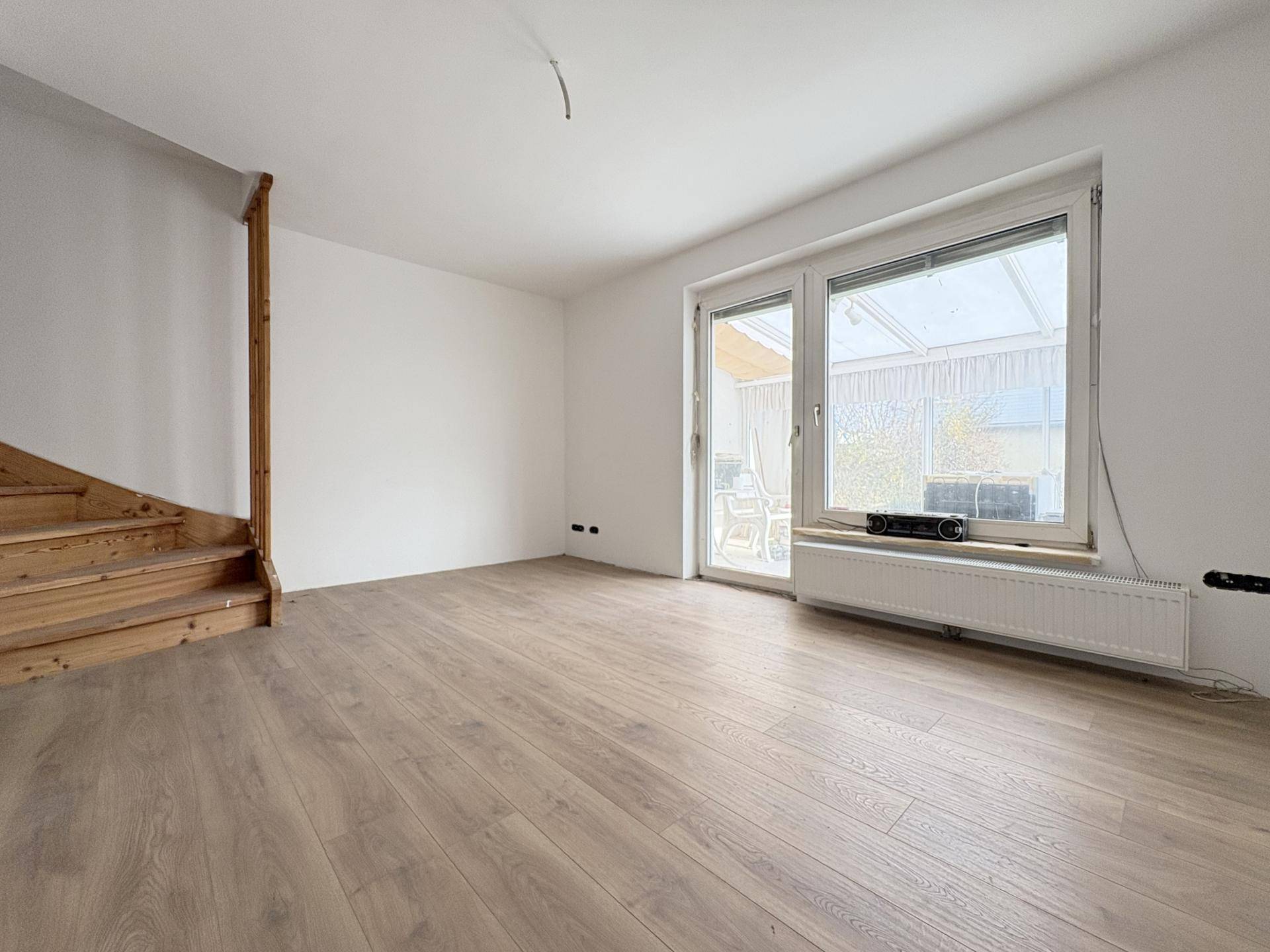 Leeres Wohnzimmer mit Laminatboden, Holztreppe und Zugang zum Wintergarten.