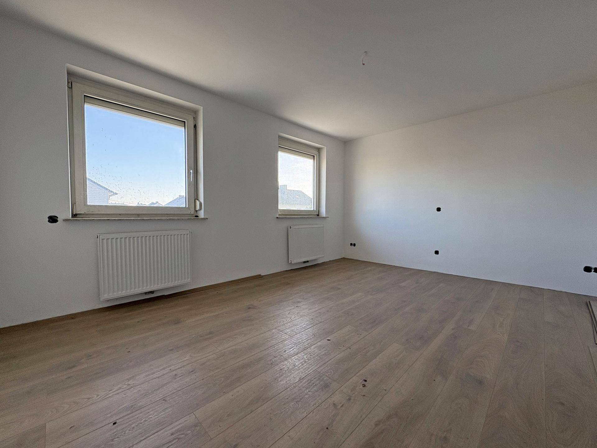 Leerer Raum mit Laminatboden, zwei Fenstern und Heizkörpern, bereit für die Gestaltung.