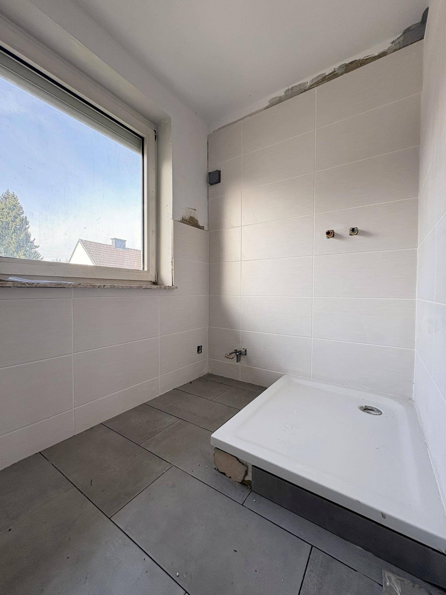 Badezimmer im Rohbau mit Duschwanne, weißen Fliesen und Fenster, bereit für die Installation.