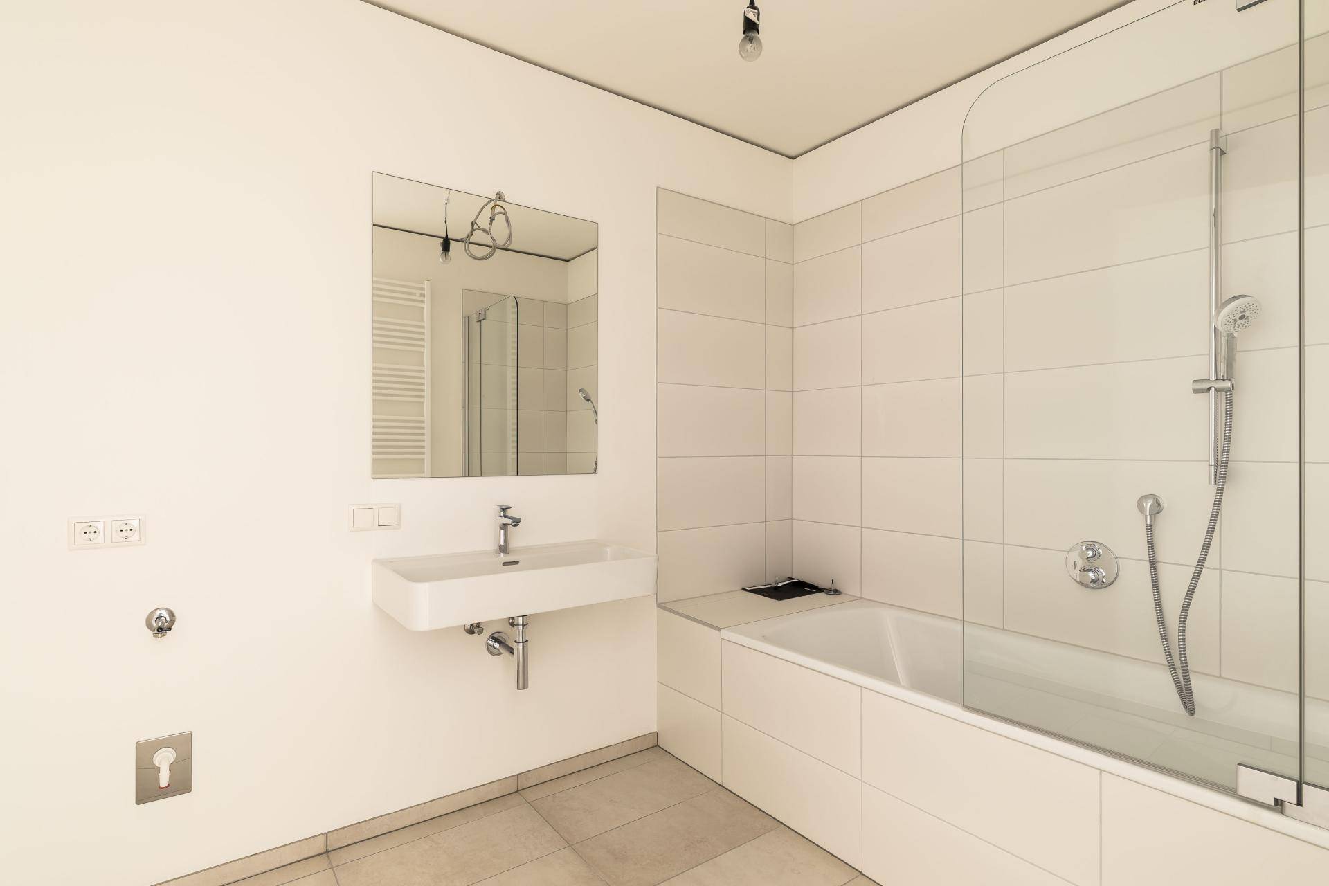 Modernes Badezimmer mit weißen Fliesen, Badewanne mit Glasabtrennung und Wandwaschbecken.