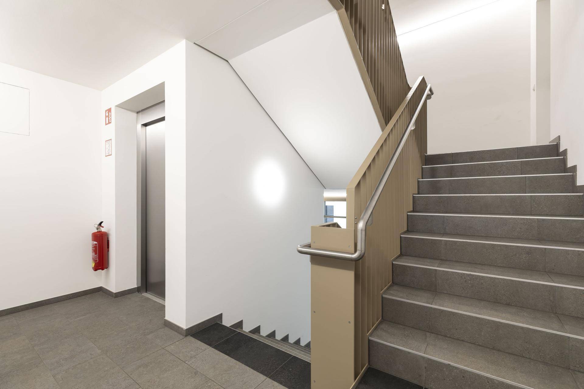 Helles Treppenhaus mit Aufzug und modernen Fliesen, ausgestattet mit einem Feuerlöscher.