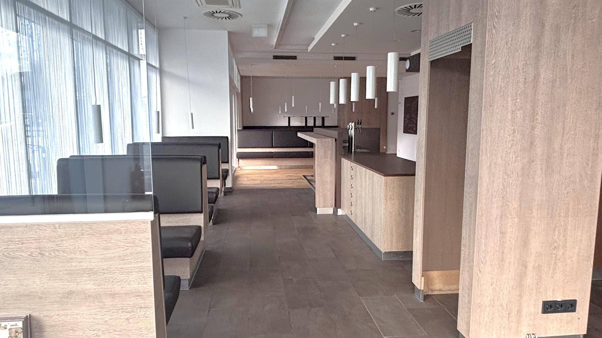 Heller Gewerberaum mit Holzelementen, Fliesenboden und bequemen Sitzbänken entlang der Fensterfront.