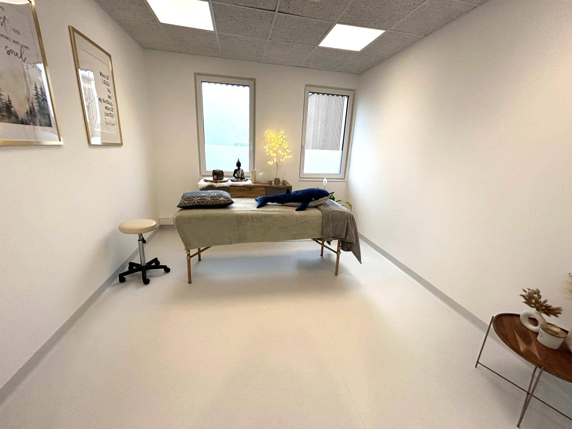 Heller Behandlungsraum mit Massageliege, großen Fenstern und minimalistischer Dekoration.