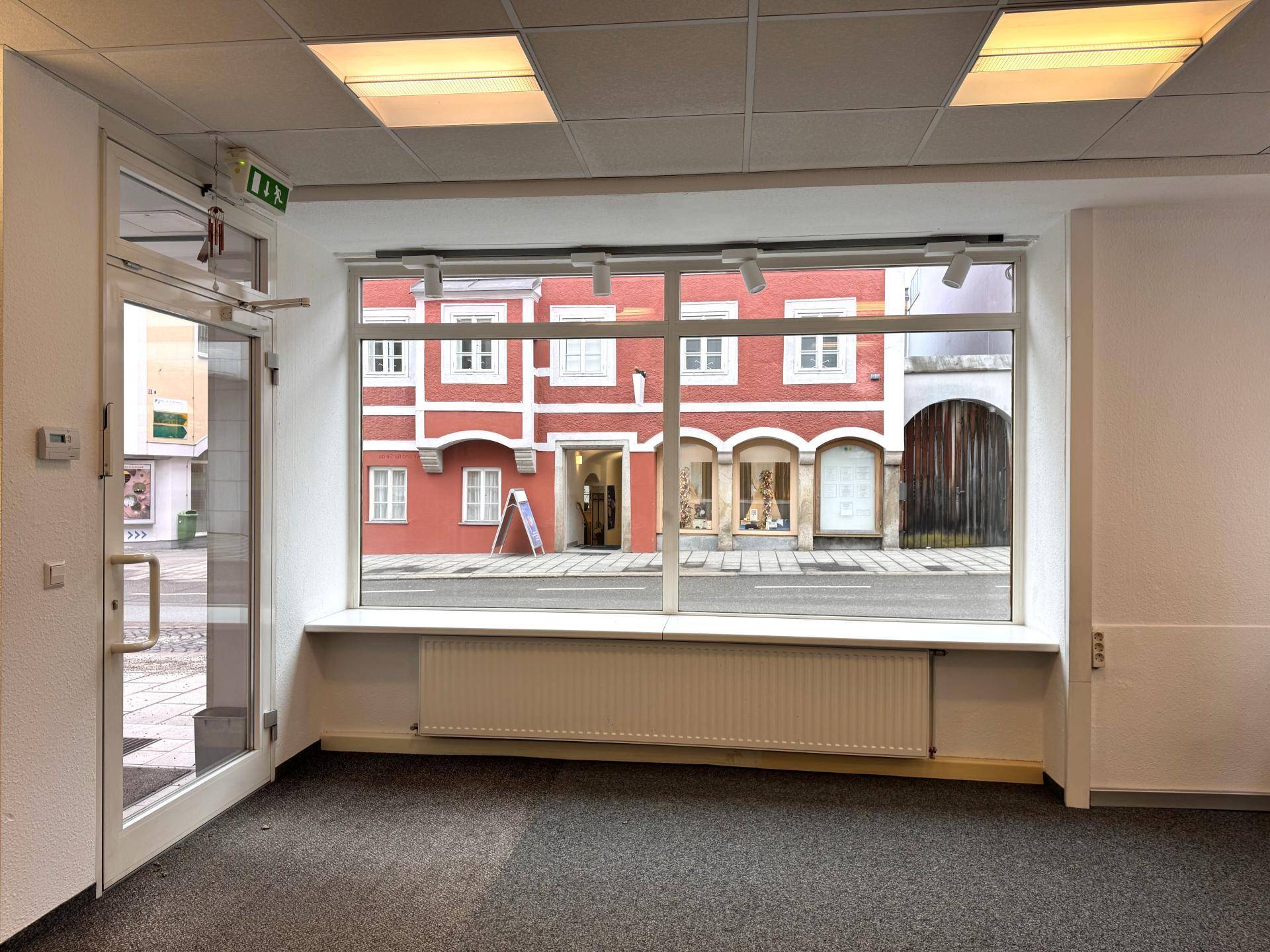 Innenansicht eines leeren Geschäftsraums mit großen Schaufenstern, die viel Tageslicht hereinlassen und Blick auf die Straße bieten.