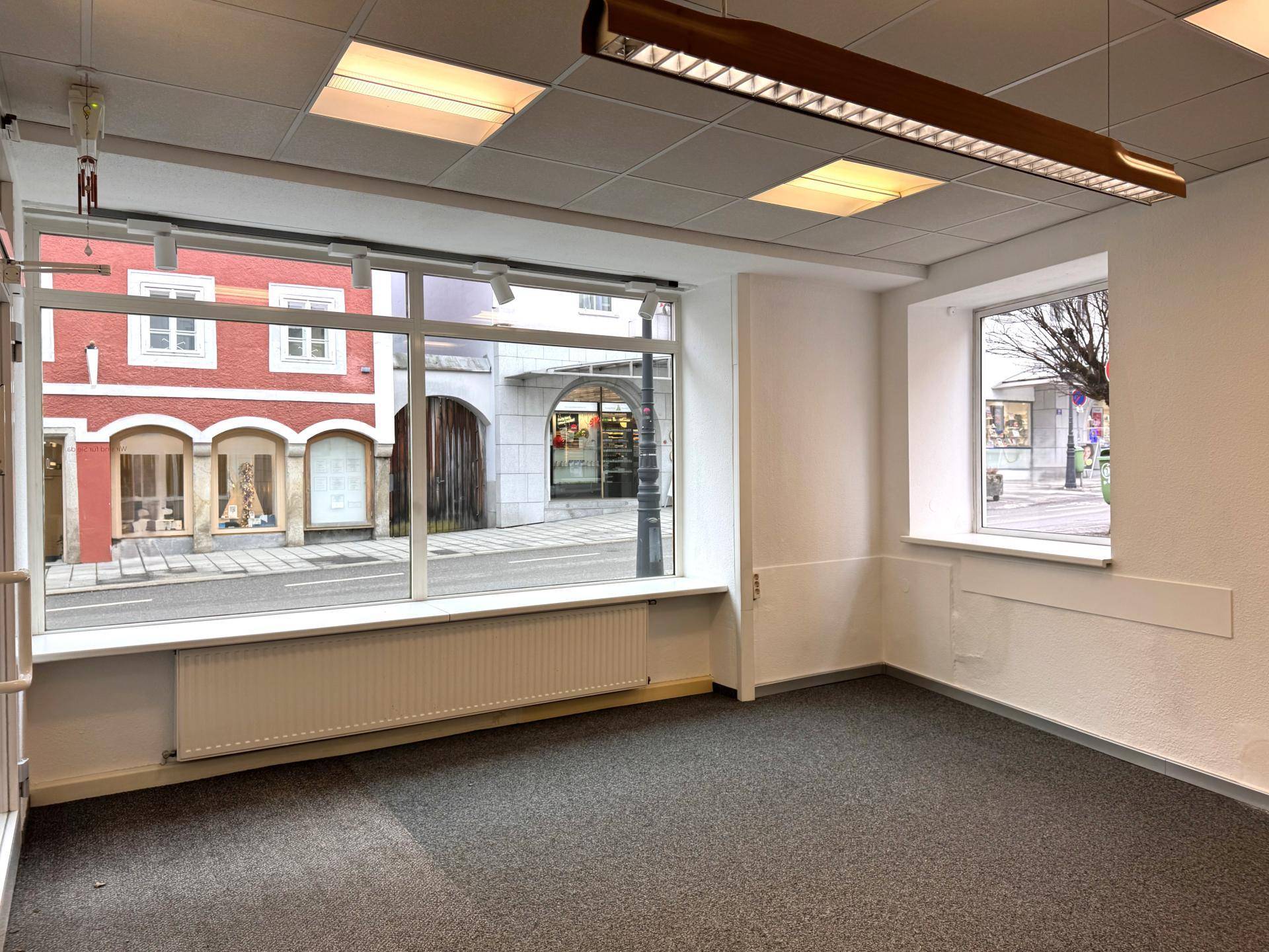 Geräumiger, leerer Geschäftsraum mit großen Fenstern zur Straße und einem kleineren Seitenfenster, ausgestattet mit Teppichboden.