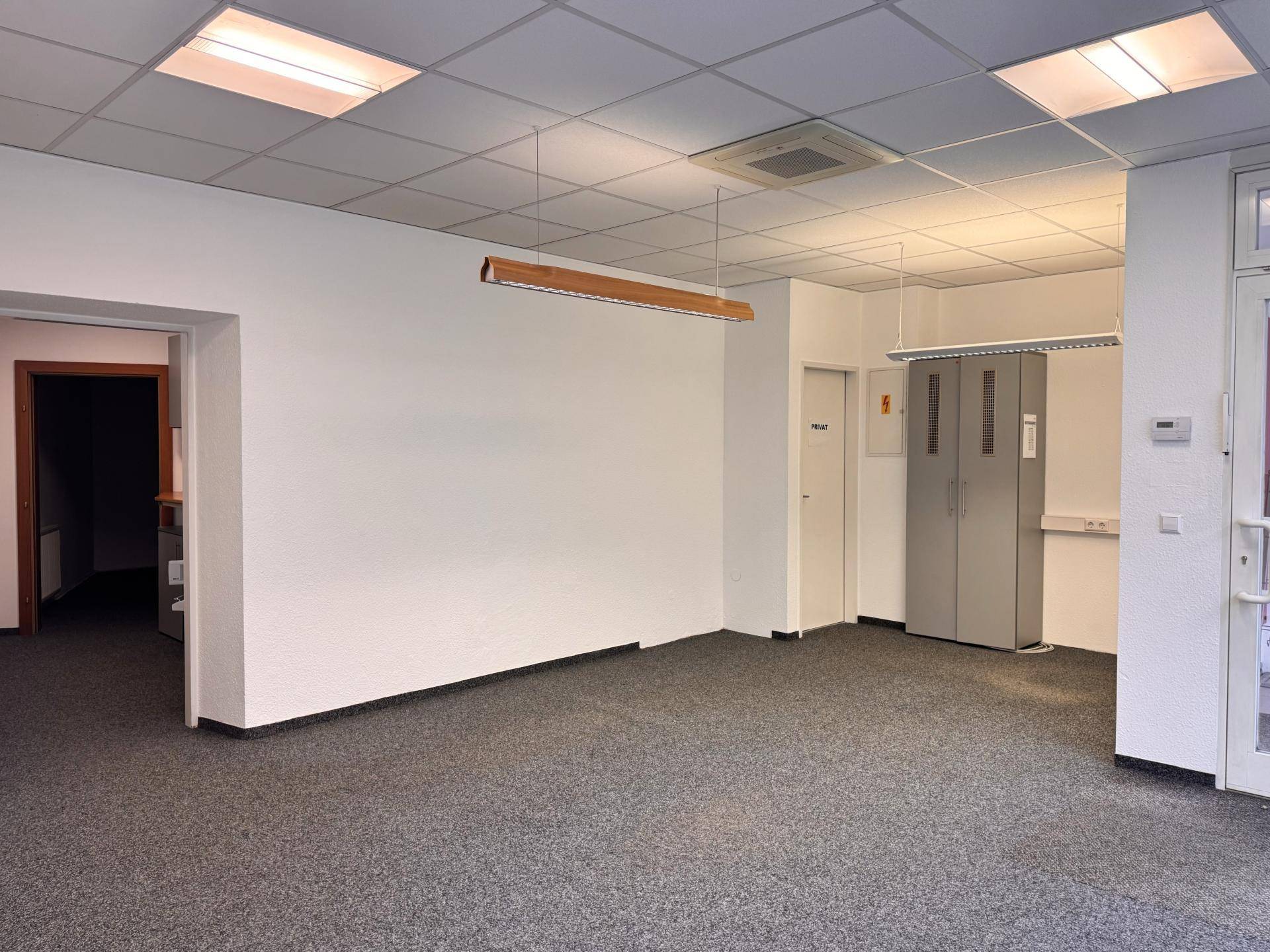 Innenansicht eines Geschäftsraums mit grauem Teppichboden, abgehängter Decke und Zugang zu weiteren Bereichen.