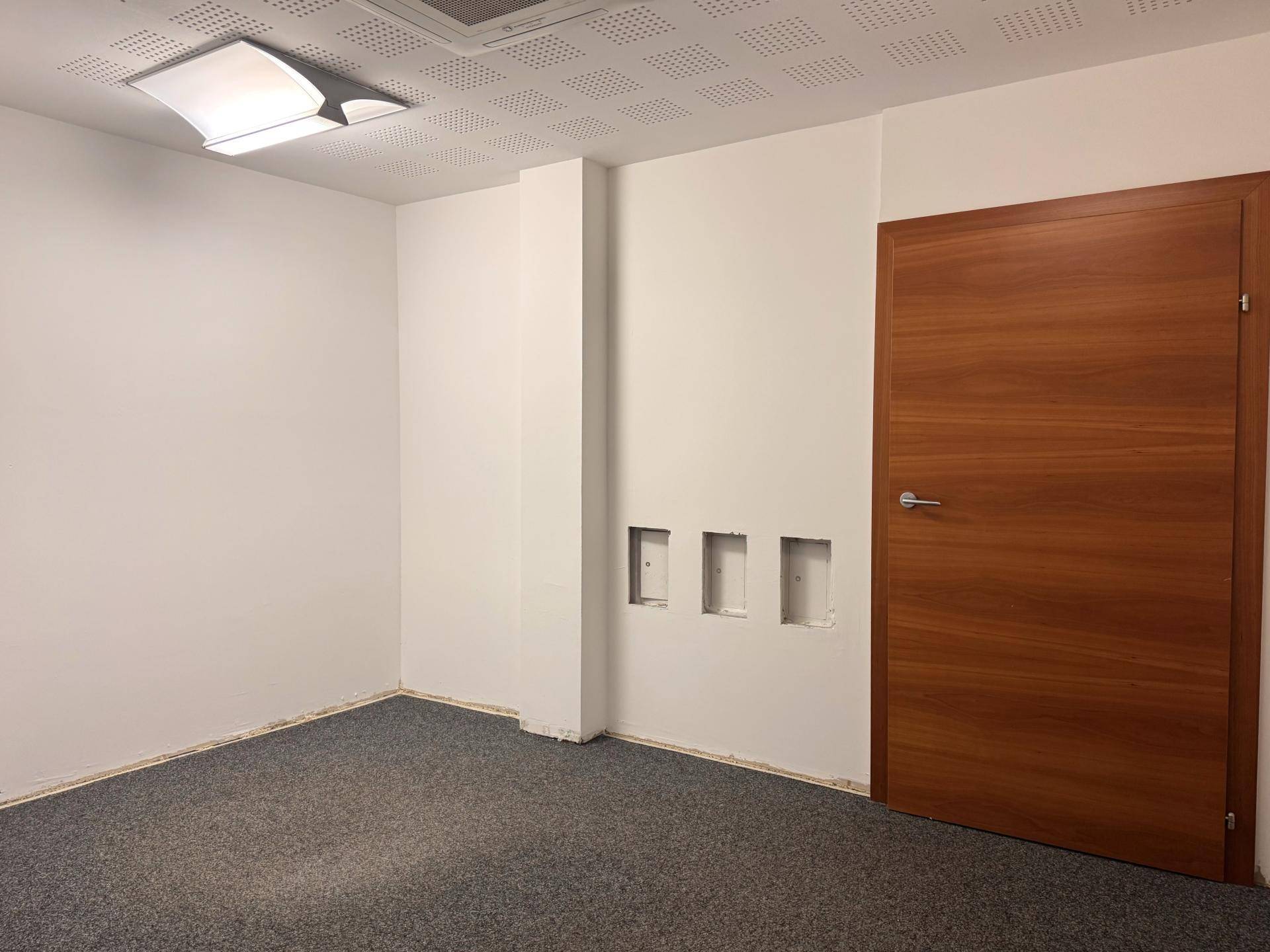 Leerer Büroraum mit weißen Wänden, grauem Teppichboden und einer modernen Holztür.