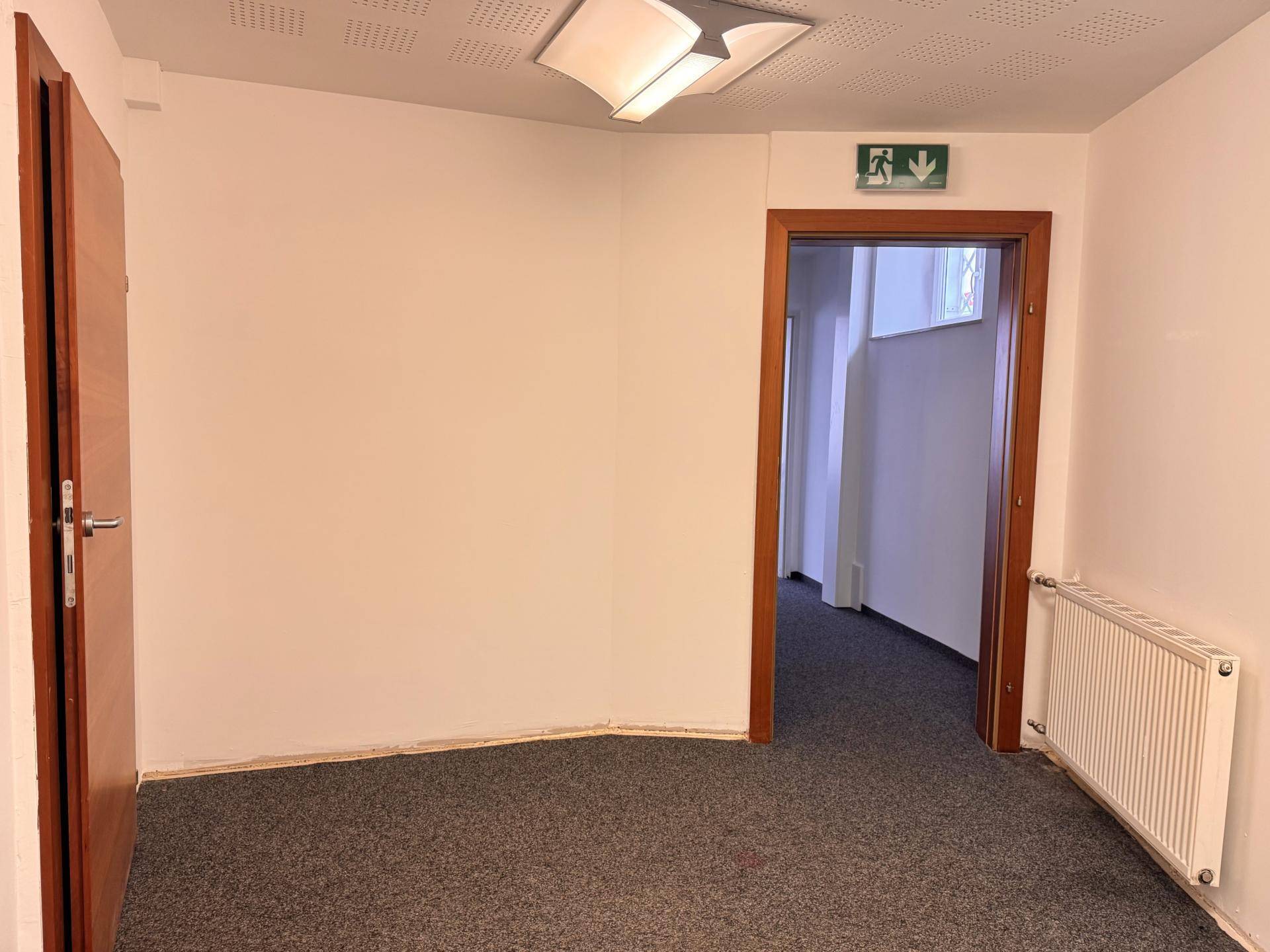 Schlichter Büroraum mit Teppichboden, Heizkörper und einer Holztür, die zu einem Flur führt.