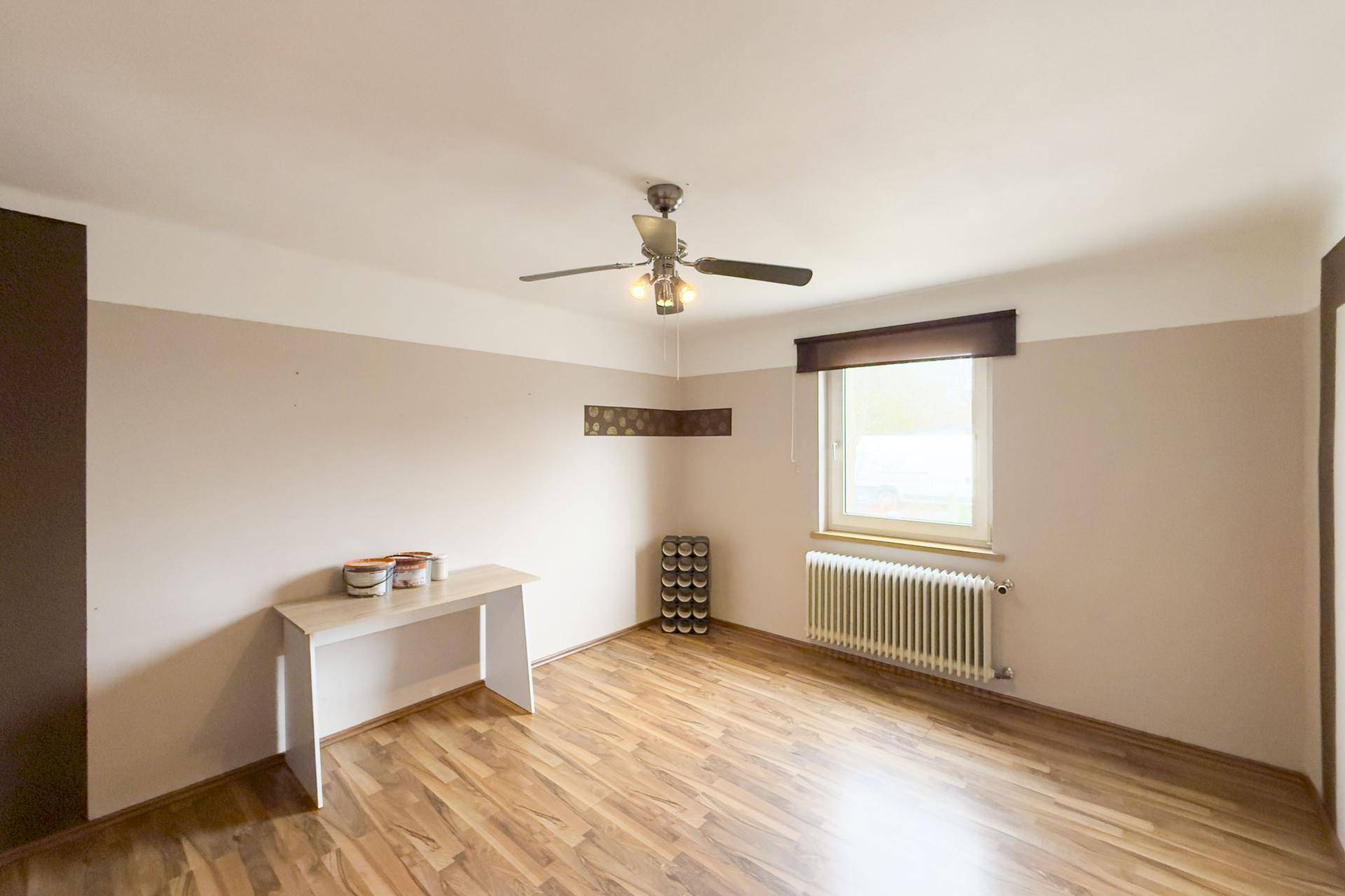 Heller Raum mit Laminatboden, Deckenventilator und Heizkörper unter dem Fenster.