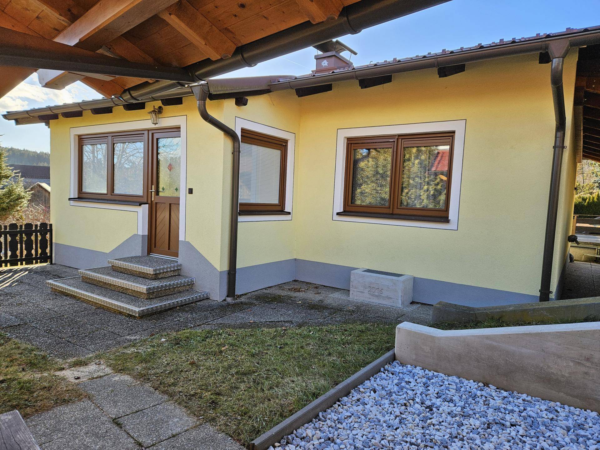 Seitenansicht des Hauses mit Eingangsbereich, braunen Fenstern und kleiner gepflasterter Fläche.