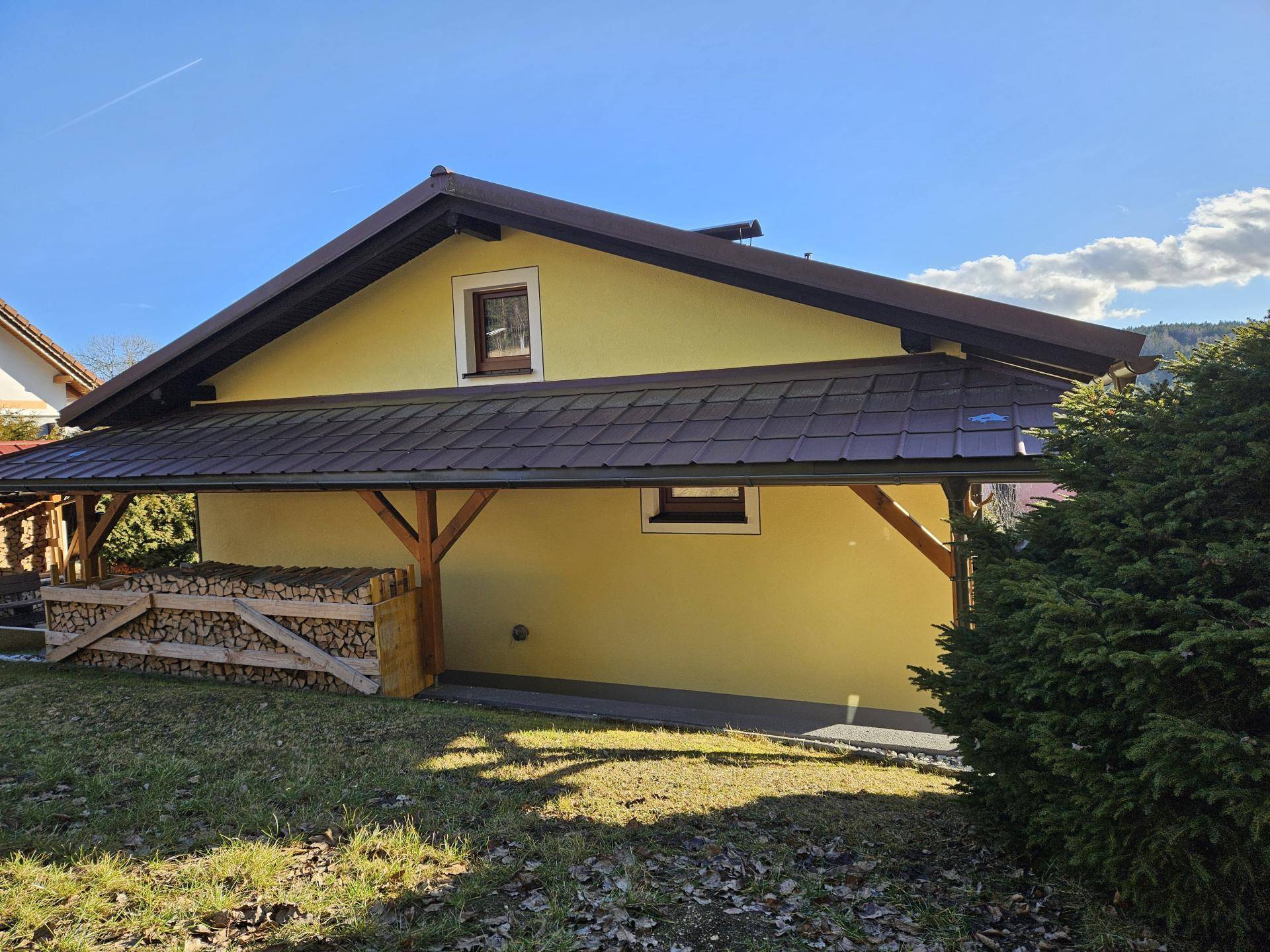 Seitenansicht des Hauses mit überdachtem Bereich, gestapeltem Brennholz und Gartenfläche.