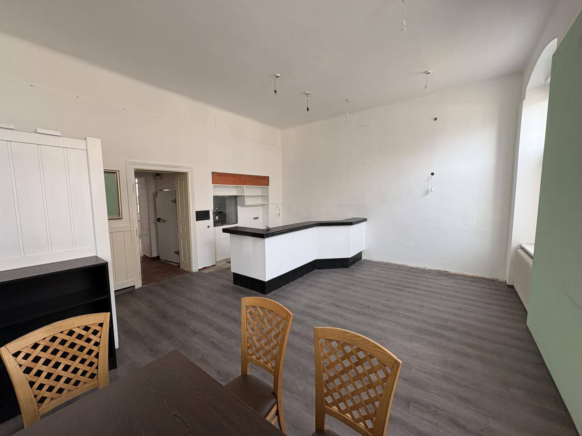 Innenraum mit einer Bar, dunklem Boden und weißen Wänden, mit Blick auf einen angrenzenden Bereich und Möbel im Vordergrund.