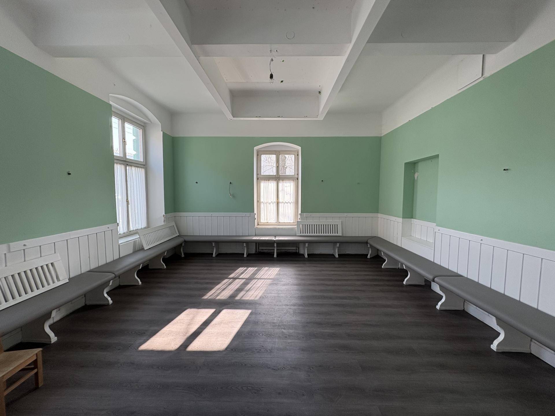 Großer, leerer Raum mit hellgrünen Wänden, dunklem Boden und Sitzbänken entlang der Wände, durchflutet von Sonnenlicht.