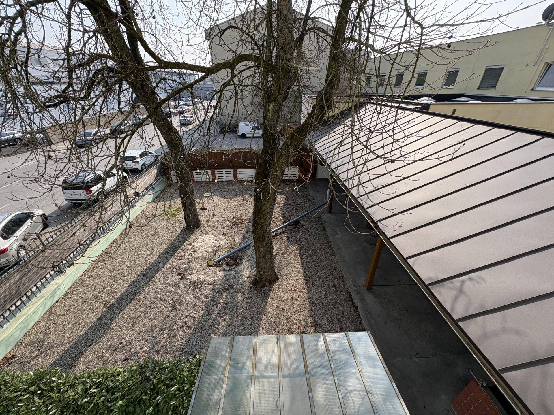 Blick von oben auf einen Außenbereich mit Kiesboden, kahlen Bäumen und einer überdachten Struktur, mit parkenden Autos im Hintergrund.