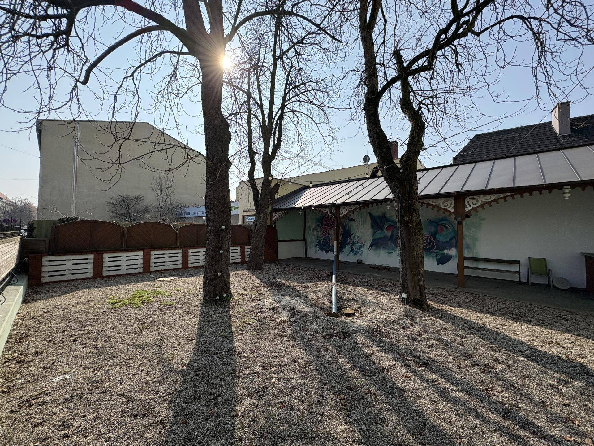 Gartenbereich mit Kiesboden, Bäumen und einer Wand mit Wandmalereien unter einem überdachten Abschnitt, bei Sonnenschein.
