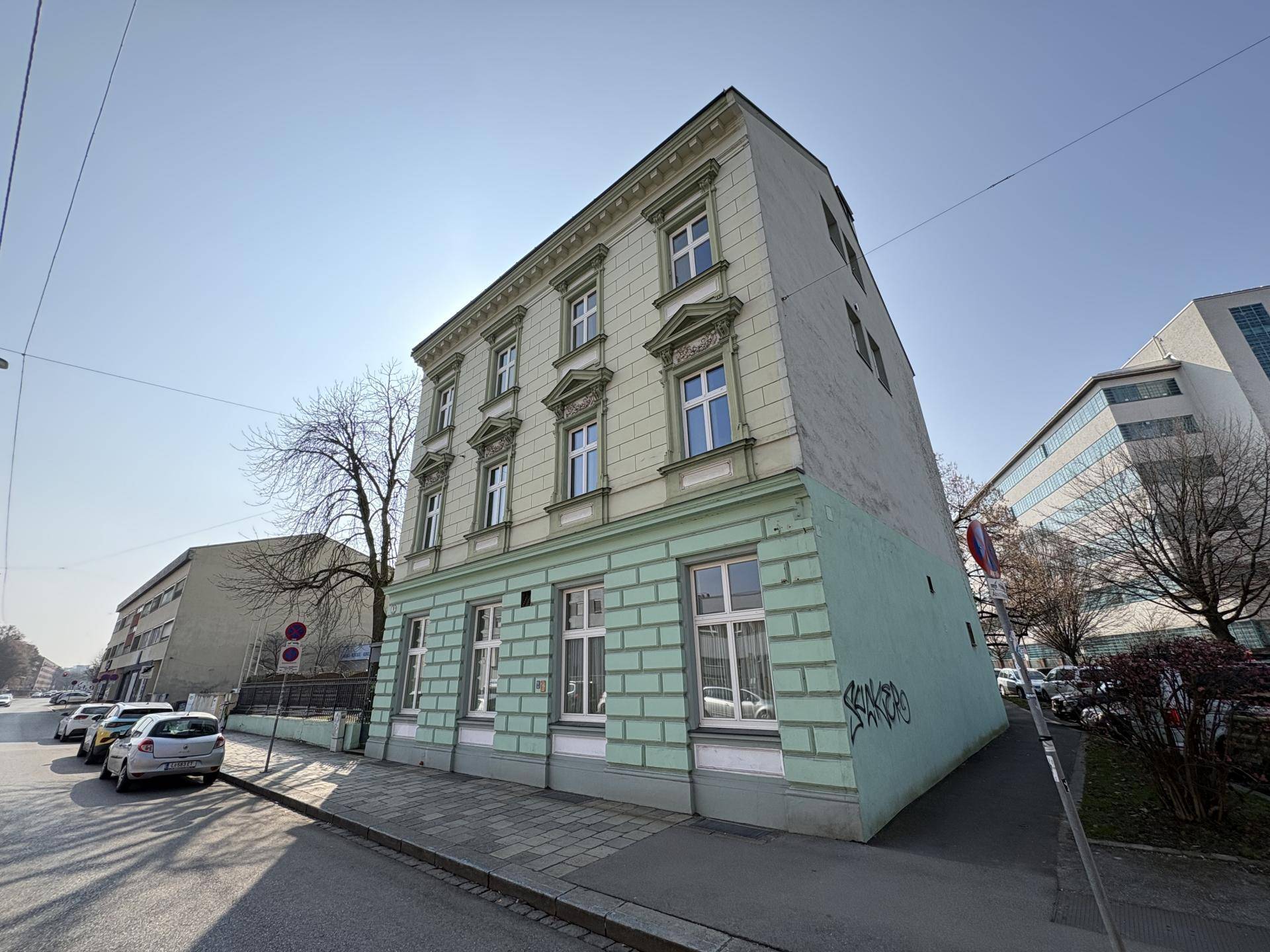 Historisches Gebäude mit hellgrüner und beiger Fassade an einer Straße mit geparkten Autos unter blauem Himmel.